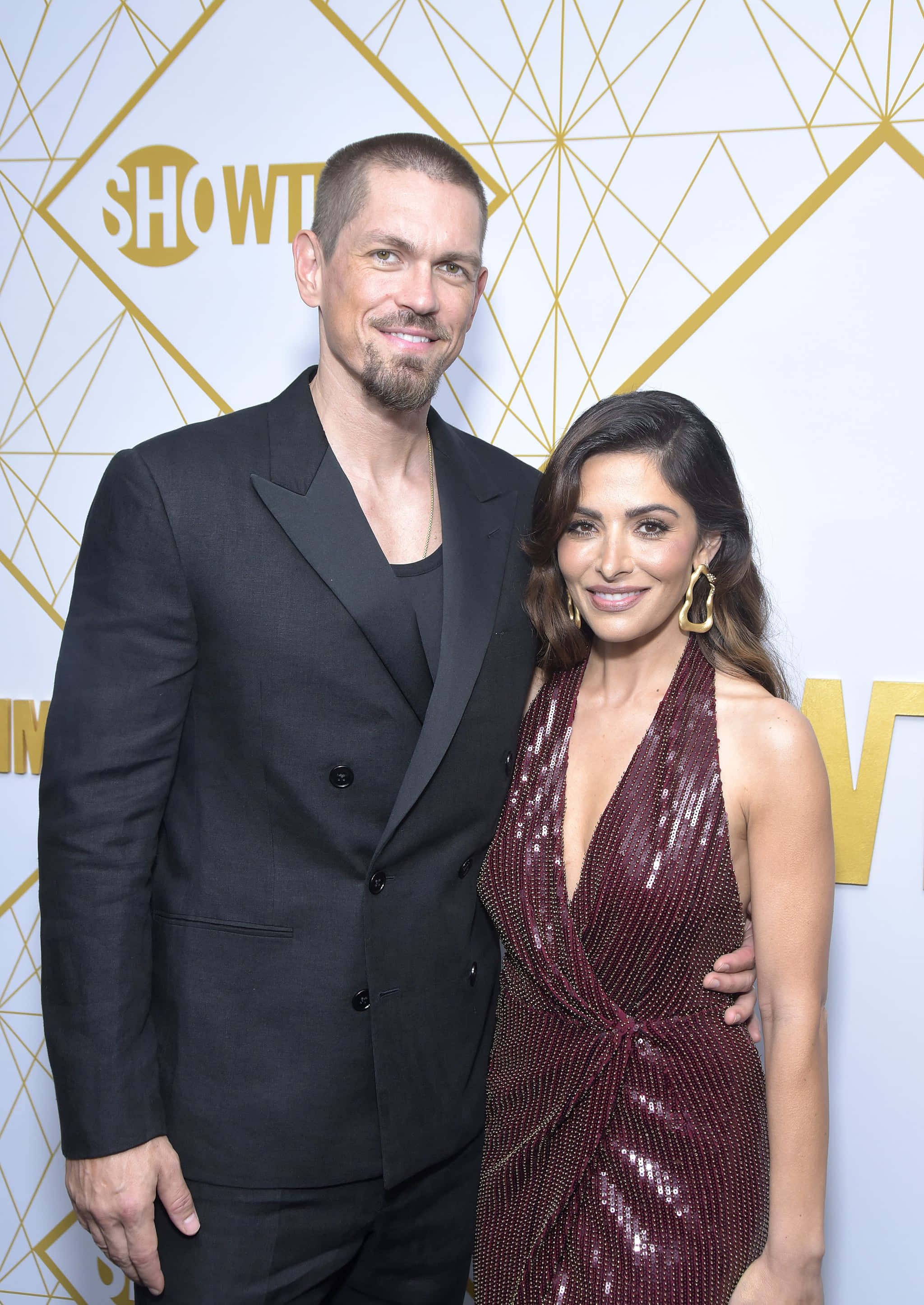 sarah shahi 桌布 壁紙 — 一名身穿黑色西裝的男子和一名身穿閃亮栗色連衣裙的女子在一個華麗的活動中，以幾何金色背景一起擺姿勢。