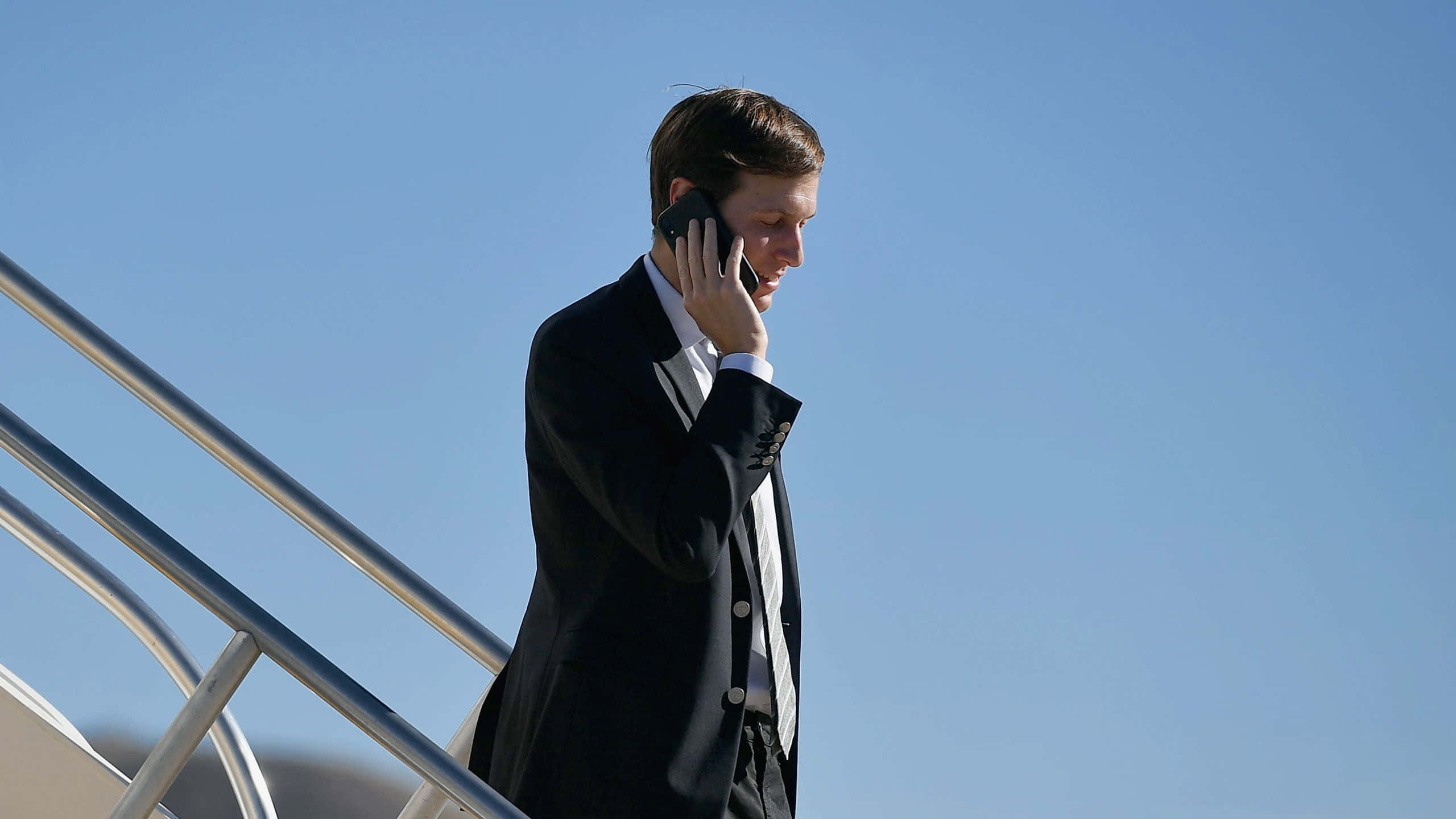 United States 桌布 壁紙 — 一名身穿黑色西裝的男子在晴朗的藍天下，一邊講電話一邊走下飛機樓梯。