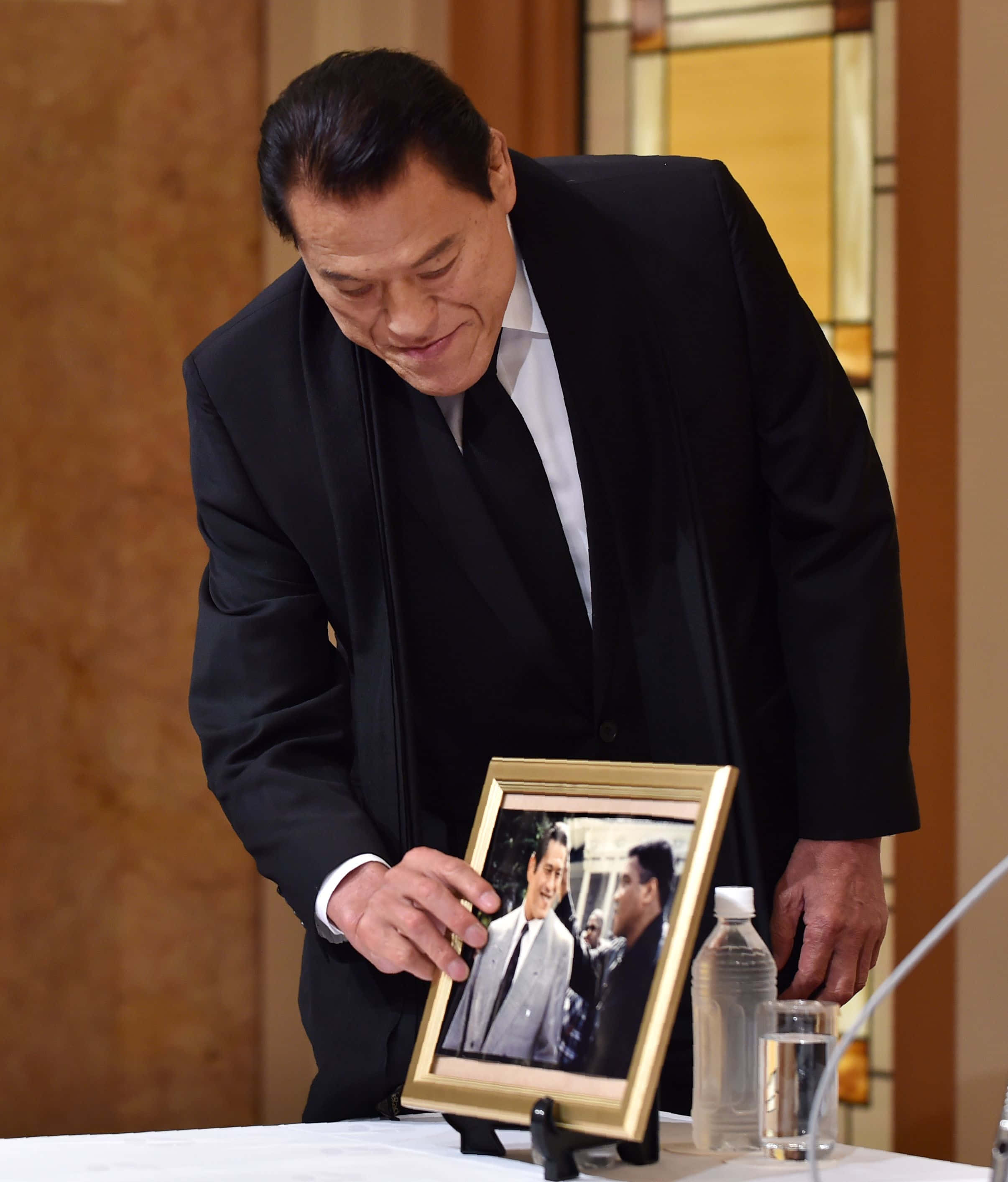 運動 桌布 壁紙 — 一名身穿黑色西裝的男子在溫暖燈光的室內環境中，輕輕觸摸桌上水瓶旁邊的一個裱框照片。