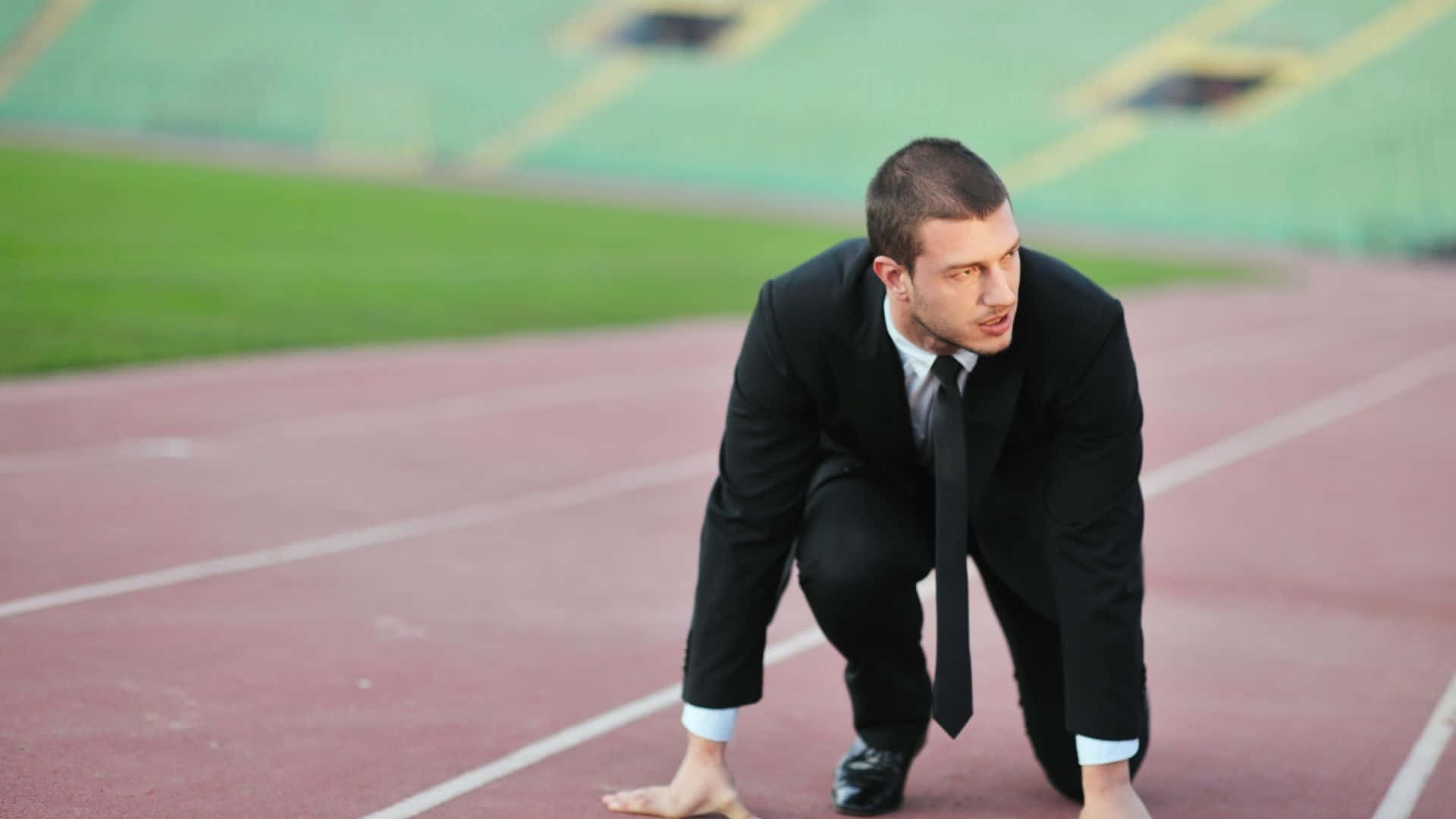 Best Sports 桌布 壁紙 — 一名身穿黑色西裝的男子在紅色跑道的起跑線上蹲下，背景是綠色的體育場座位。