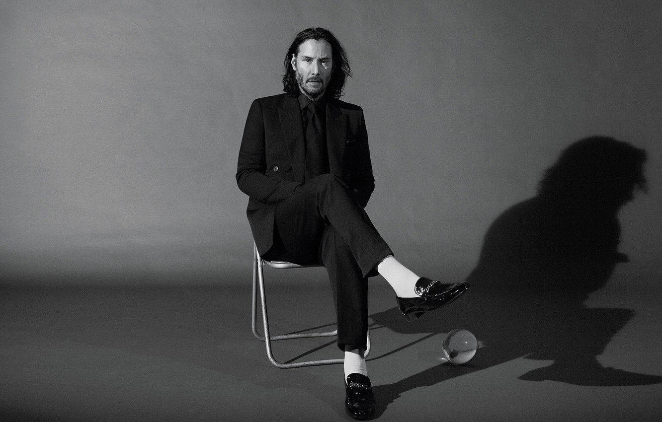 Keanu 桌布 壁紙 — 一名身穿黑色西裝的男子坐在椅子上，在灰色背景上投射出戲劇性的陰影，腳邊有一個玻璃球體。