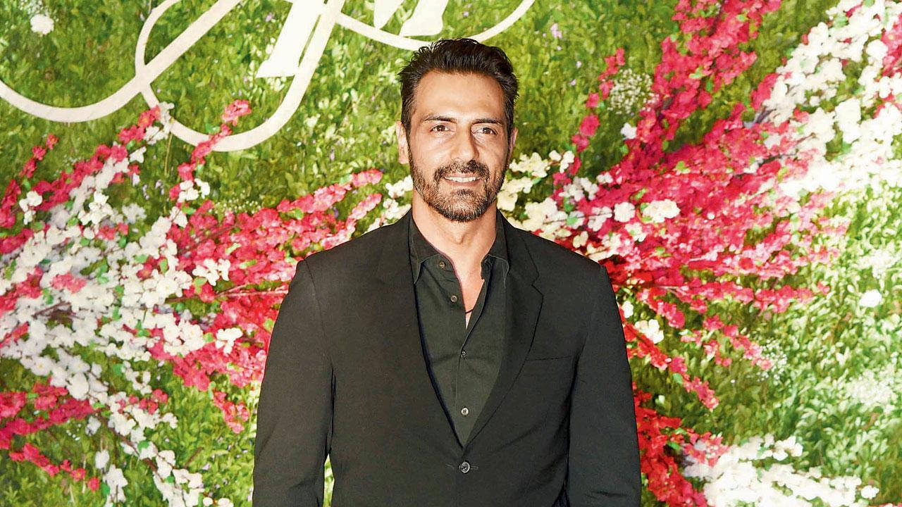 Arjun Rampal 桌布 壁紙 — 一名身穿黑色西裝的男子自信地站在一面充滿粉紅色、白色和綠色花朵的茂盛花牆前。