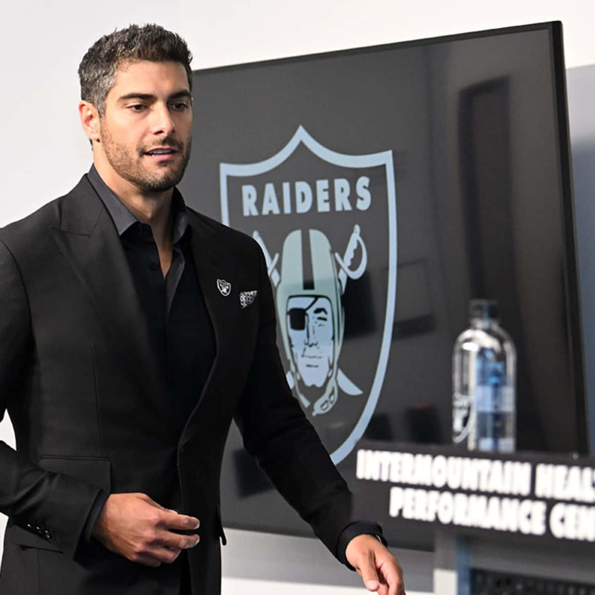 Jimmy Garoppolo 桌布 壁紙 — 一名身穿黑色西裝的男子自信地站在表演中心螢幕上的 Raiders 標誌前面。