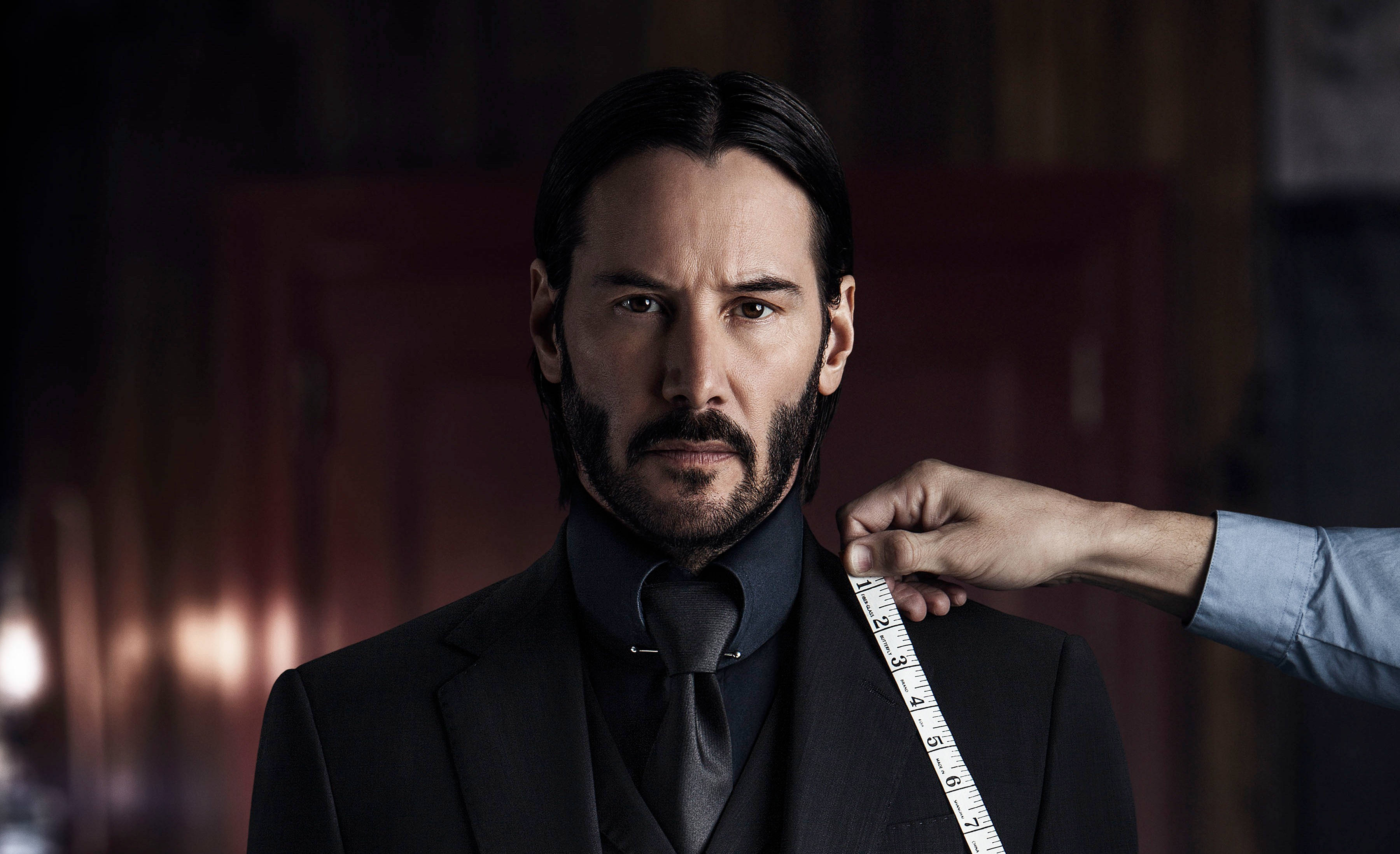 John Wick 桌布 壁紙 — 一名身穿黑色西裝的男子莊嚴地站著，一隻手在昏暗的房間裡用軟尺測量他的肩膀。