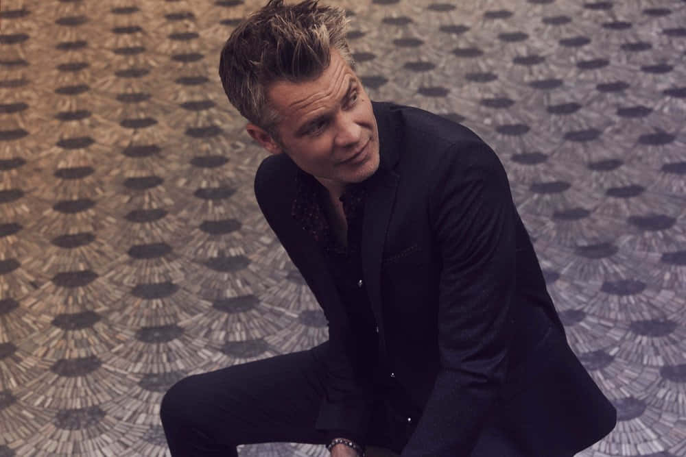 timothy olyphant 桌布 壁紙 — 一名身穿黑色西裝的男子蹲在圖案瓷磚地板上，露出自信的笑容。
