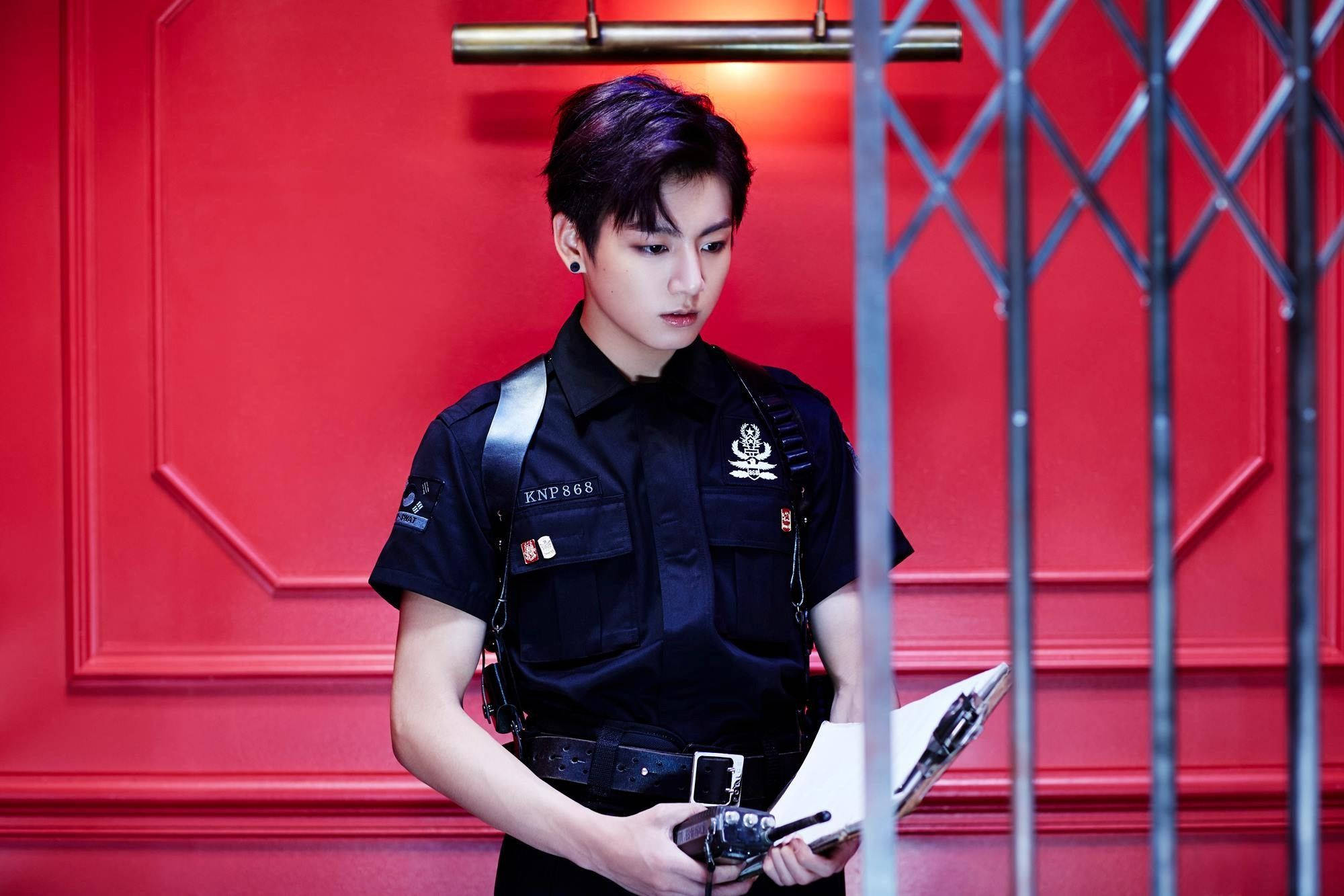 Jungkook 桌布 壁紙 — 一名身穿黑色警察制服的年輕男子站在金屬欄杆後面，背景是鮮紅色的鑲板牆，手持寫字板。