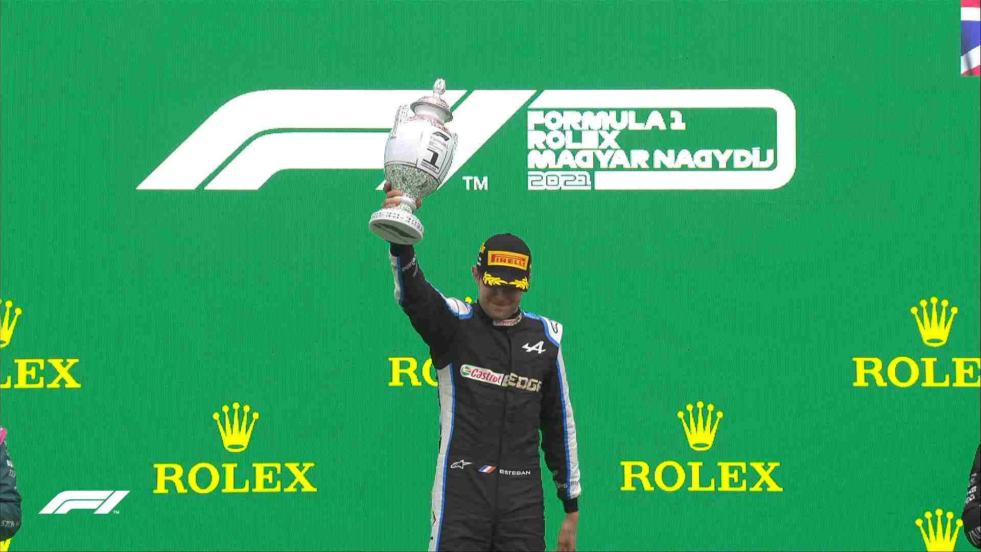 F1 桌布 壁紙 — 一名身穿黑色賽車服的F1車手在帶有Rolex和F1標誌的綠色頒獎台上，高舉銀色獎盃慶祝勝利。