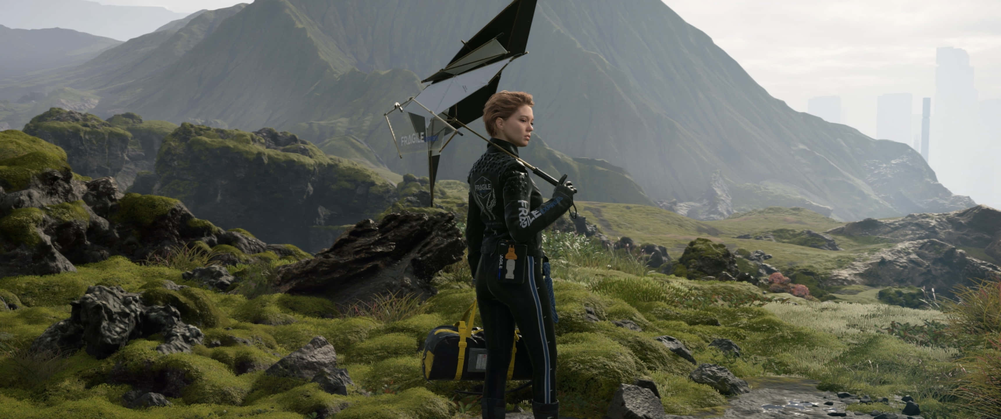 3440x1440p Death Stranding 桌布 壁紙 — 一名身穿黑色賽車服的女士站在長滿苔蘚的岩石山坡上，手持一面黑色小帆，遠處可見未來主義的城市天際線。
