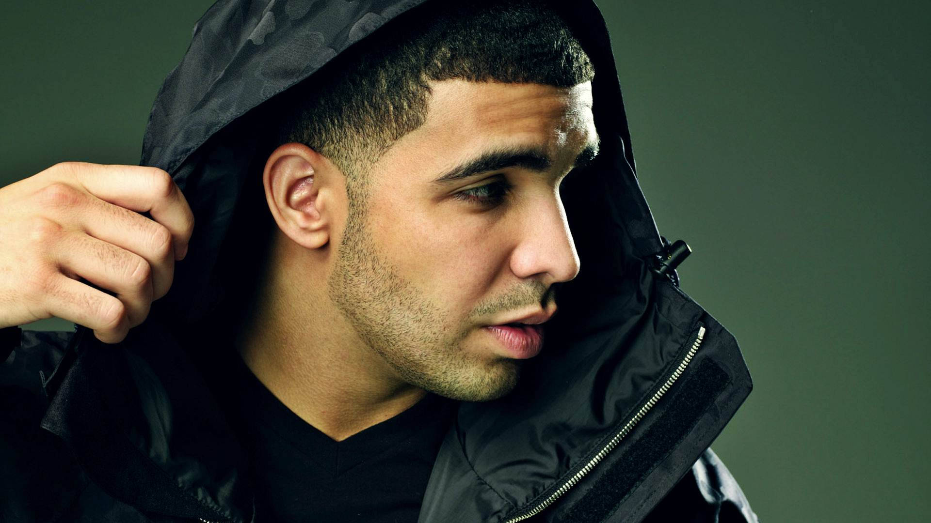 Drake 桌布 壁紙 — 一名身穿黑色連帽外套的男子拉起帽子，側頭望向一邊，背景是柔和的綠色。