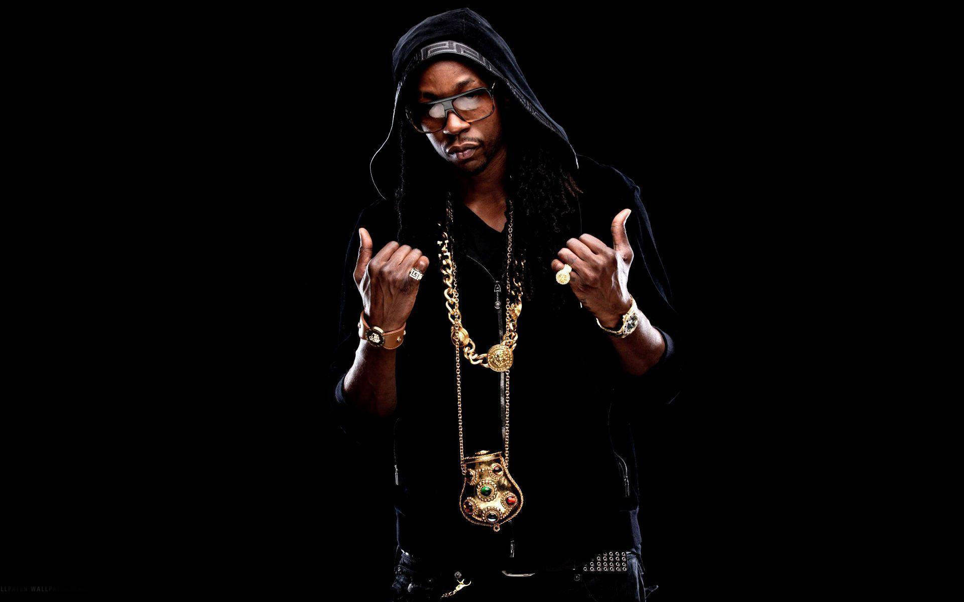 2 Chainz 桌布 壁紙 — 一名身穿黑色連帽衫、戴眼鏡的男子，在飾有金鏈和戒指的黑色背景前自信地擺姿勢。