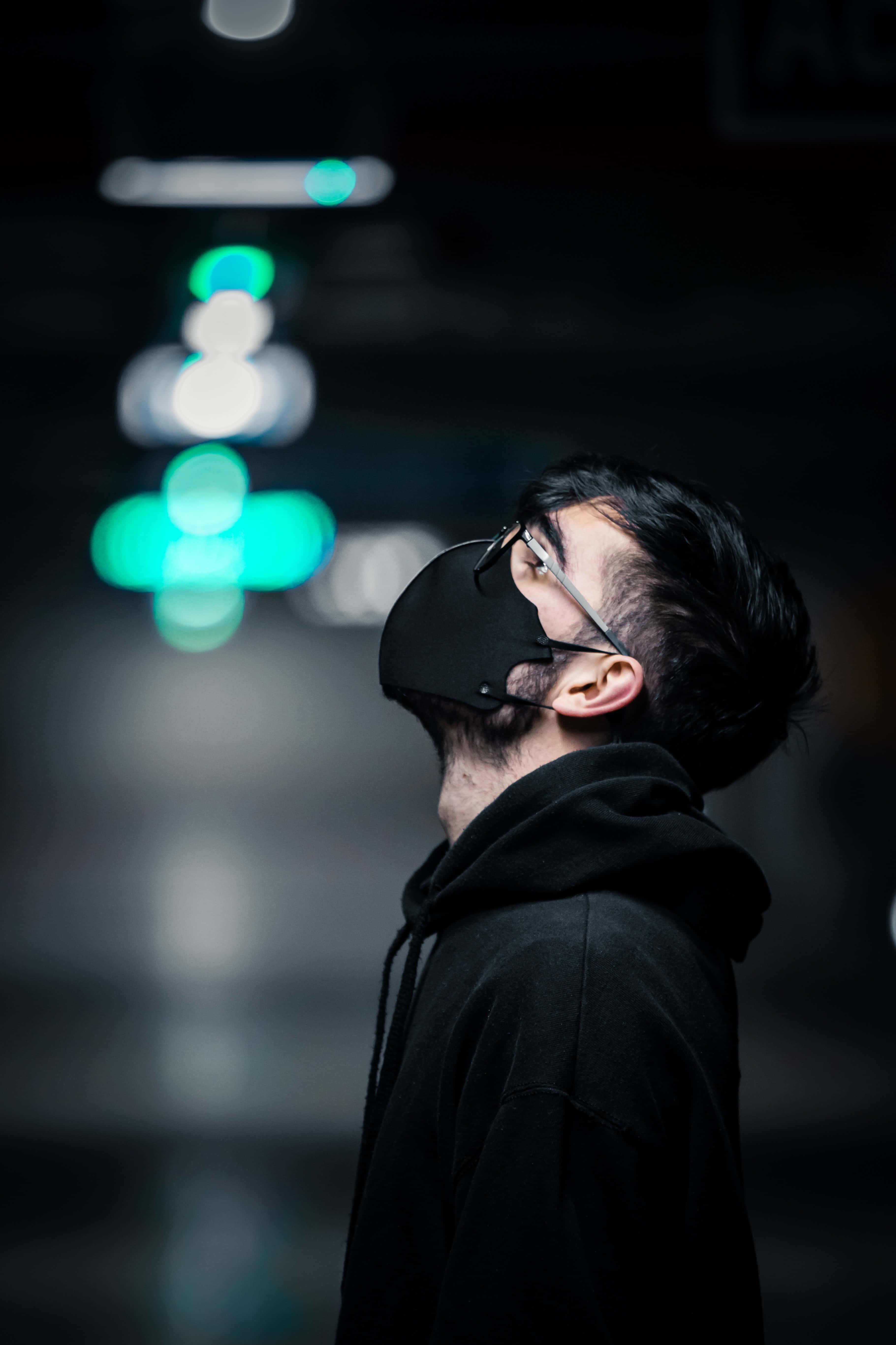 Mask 桌布 壁紙 — 一名身穿黑色連帽衫和口罩的男子在光線昏暗的城市隧道中向上凝視，周圍有發光的綠色散景燈光。