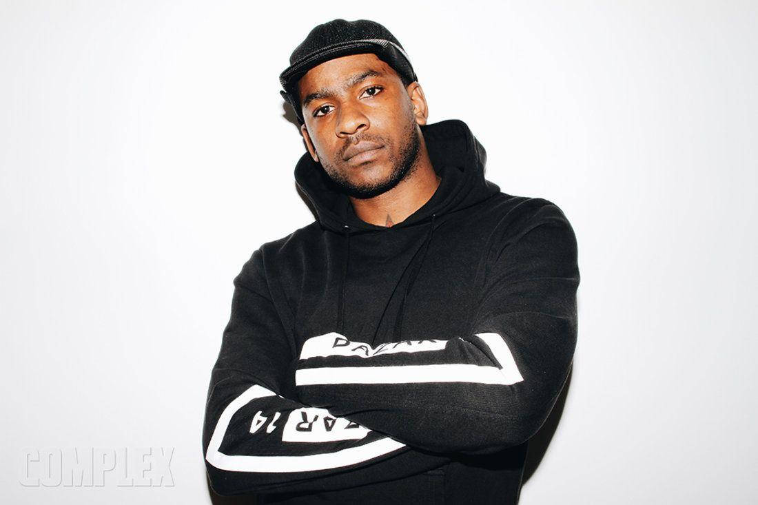 Skepta 桌布 壁紙 — 一名身穿黑色連帽衫和帽子的男子，雙臂交叉，自信地站在純白色背景前。