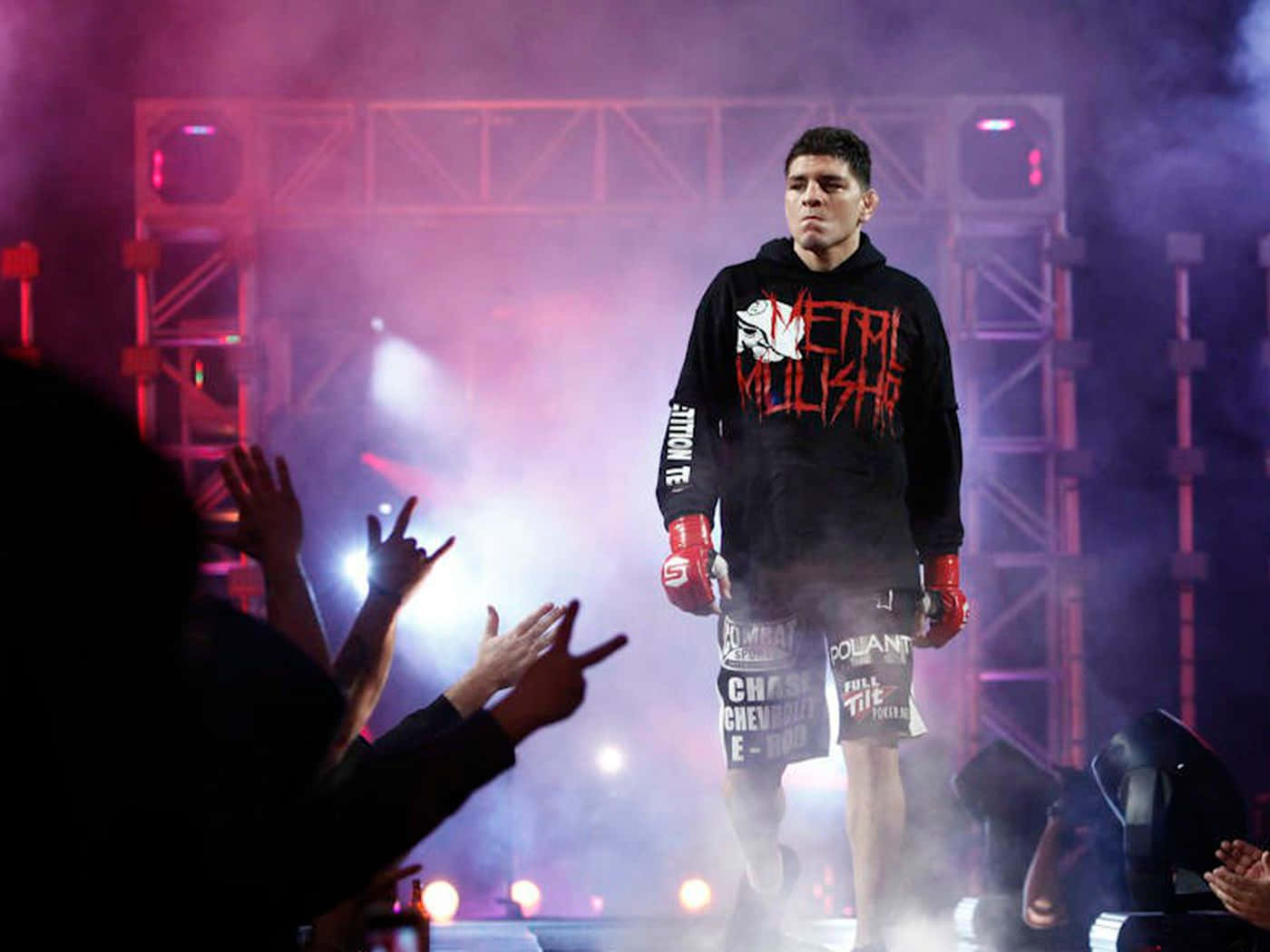 UFC 桌布 壁紙 — 一名身穿黑色連帽衫和紅色手套的戰士在舞台燈光下自信地穿過煙霧，粉絲們歡呼並舉起雙手。