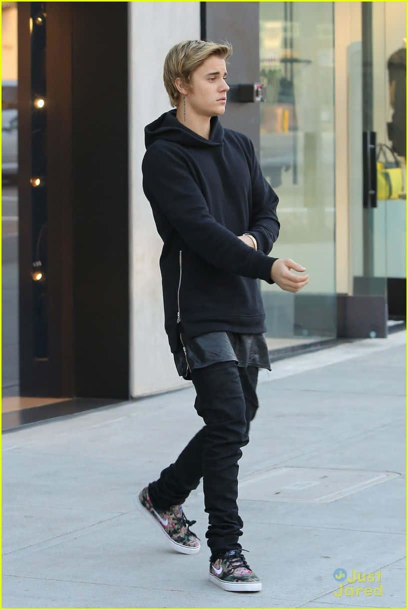 Justin Bieber 2015 桌布 壁紙 — 一名身穿黑色連帽衫和花卉運動鞋的年輕男子自信地走在城市人行道上，靠近玻璃店面。