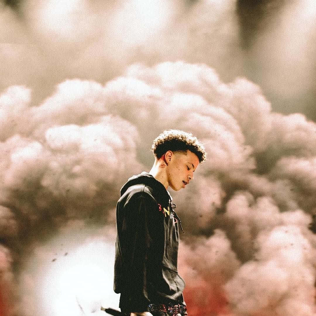 Lil Mosey 桌布 壁紙 — 一名身穿黑色連帽衫的年輕男子，在旋轉的粉紅色和灰色煙霧背景下莊嚴地站立。