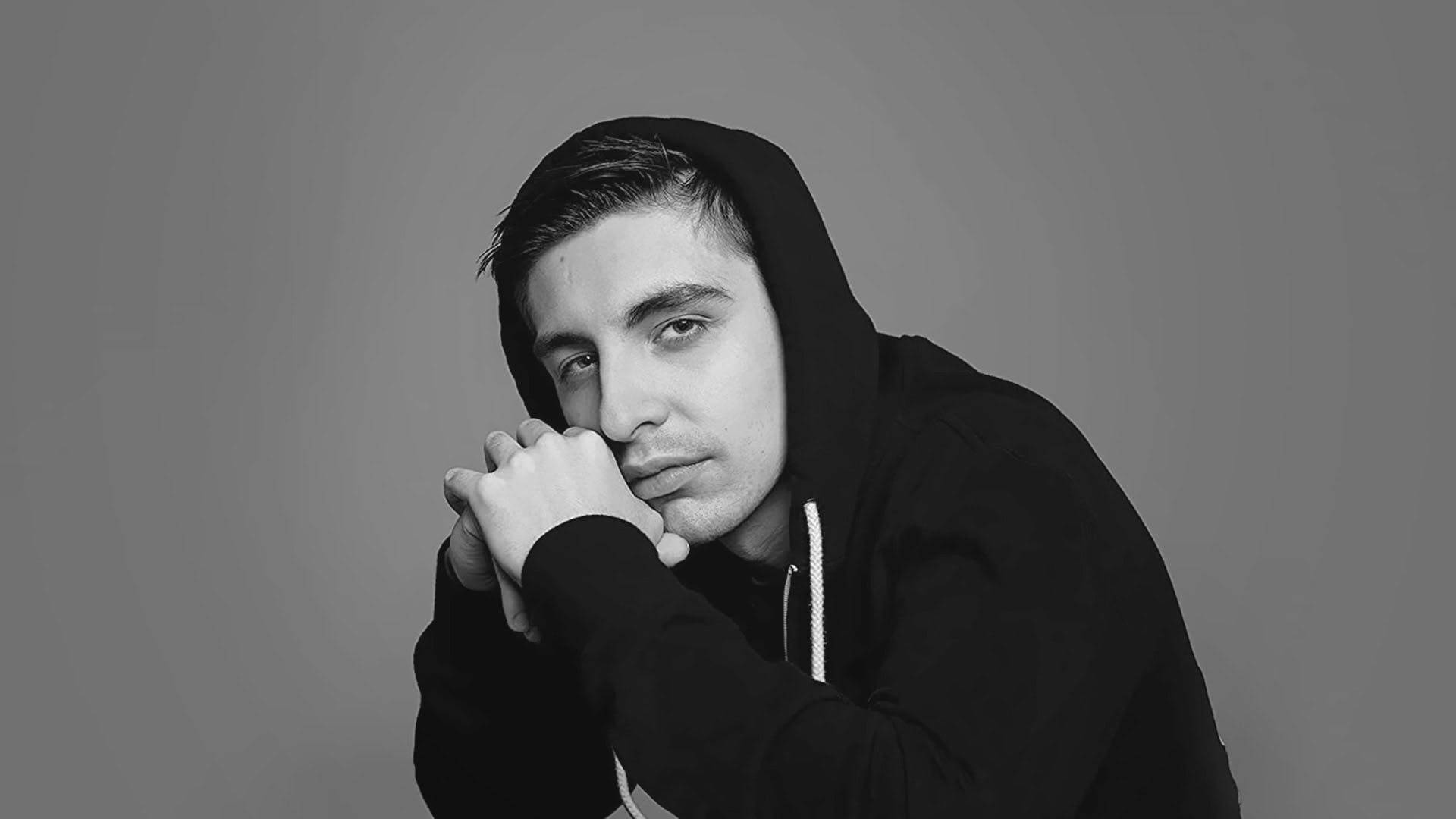 Shroud 桌布 壁紙 — 一名身穿黑色連帽衫的年輕男子，在純灰色背景前將下巴擱在手上，呈現出一幅陰鬱的黑白肖像。