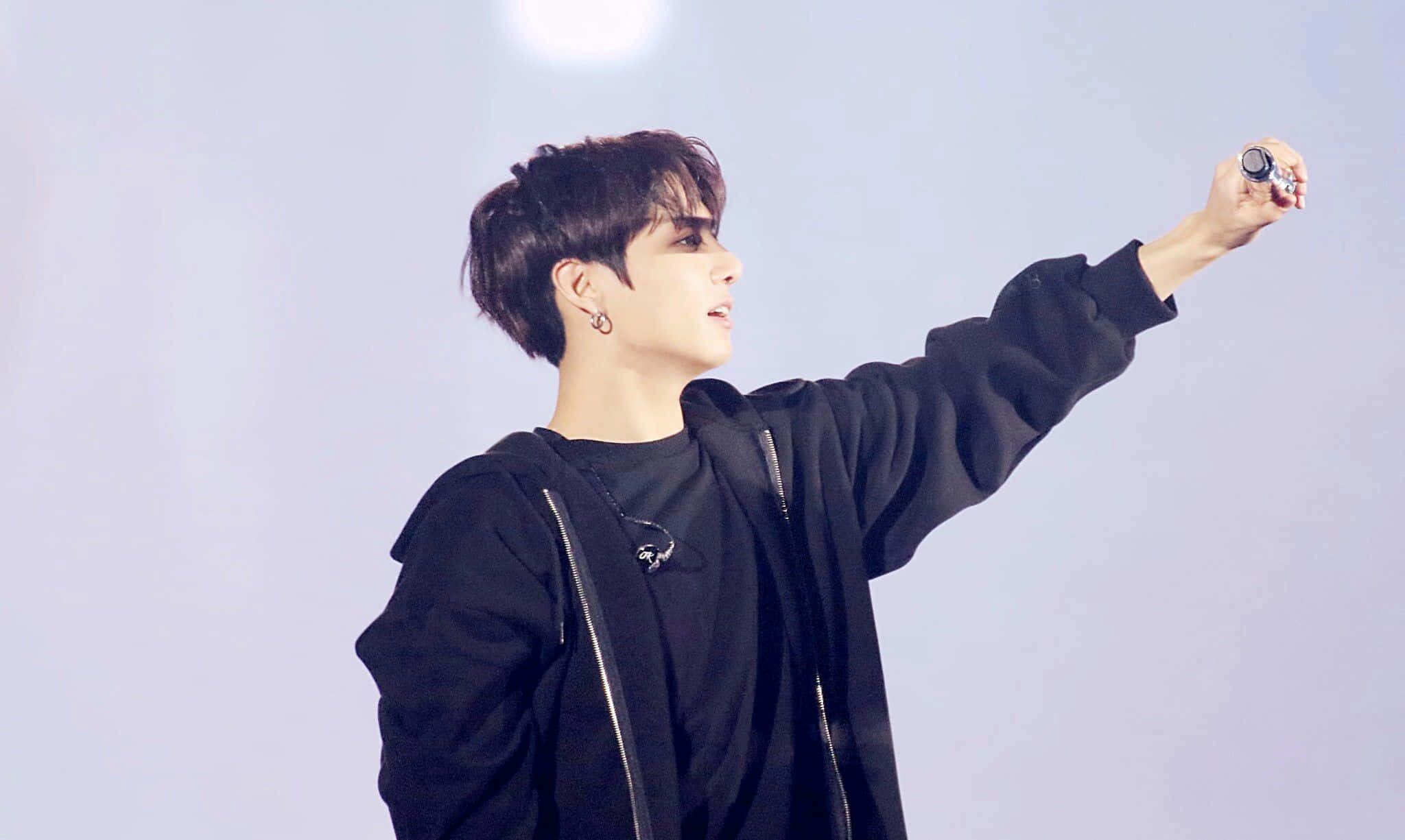 Jungkook 桌布 壁紙 — 一名身穿黑色連帽衫的年輕男子在明亮的舞台燈光下，一邊舉起麥克風，一邊向上做手勢。