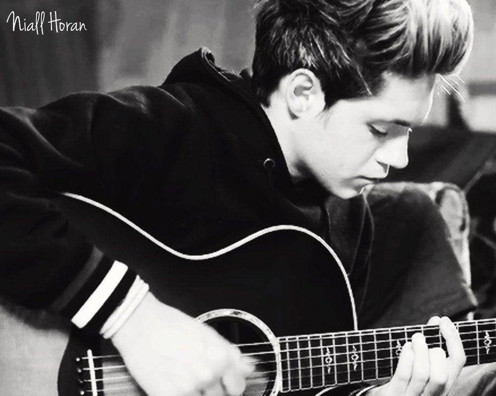 niall horan 桌布 壁紙 — 一名身穿黑色連帽衫的年輕男子在柔和光線下以單色專注地彈奏木吉他。
