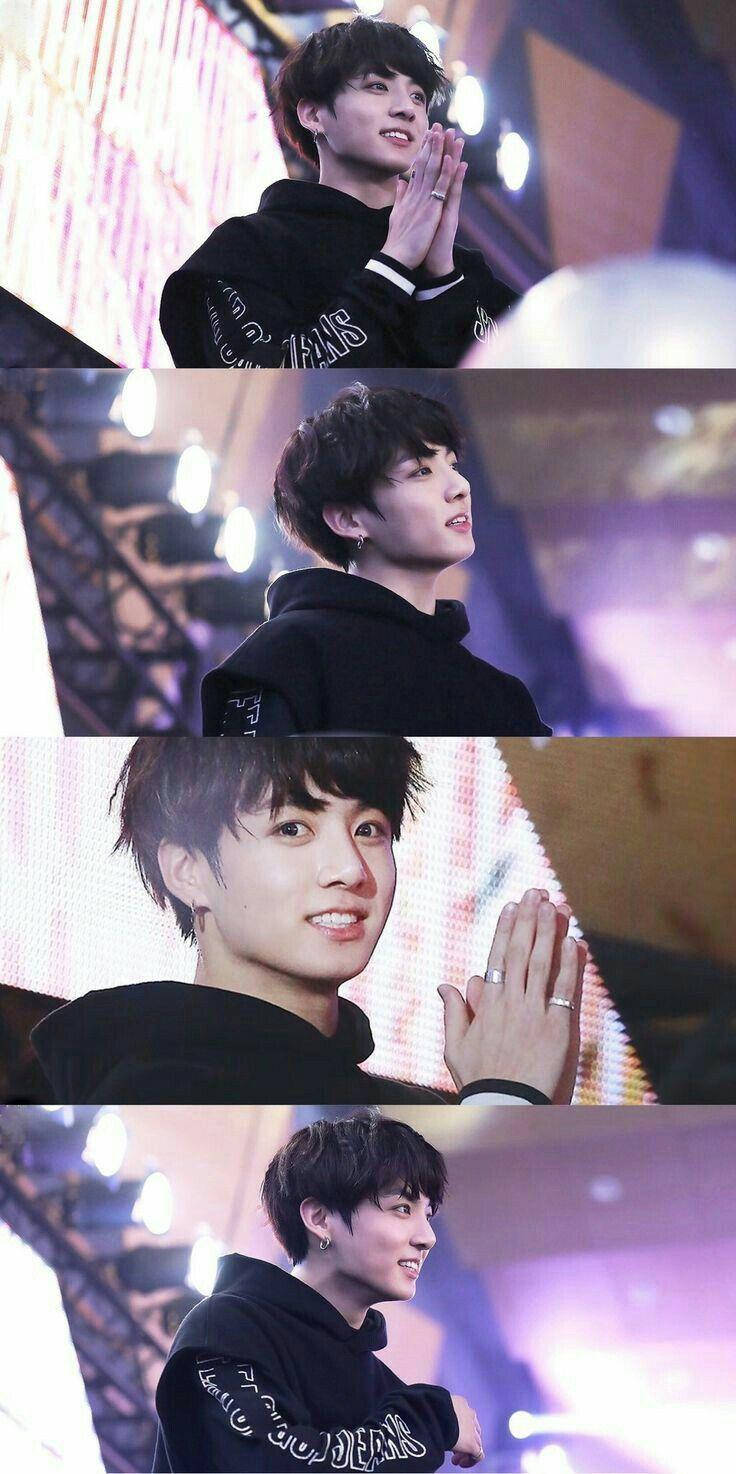 Jungkook Aesthetic 桌布 壁紙 — 一名身穿黑色連帽衫的年輕男子在燈光璀璨的舞台上微笑並鼓掌，身後是五彩繽紛的燈光和設備。
