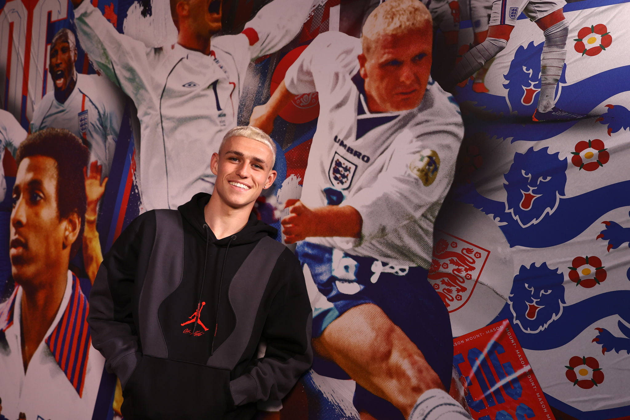 England Football 桌布 壁紙 — 一名身穿黑色連帽衫的年輕男子在英格蘭足球傳奇和國家象徵的充滿活力的牆壁拼貼畫前自信地微笑。