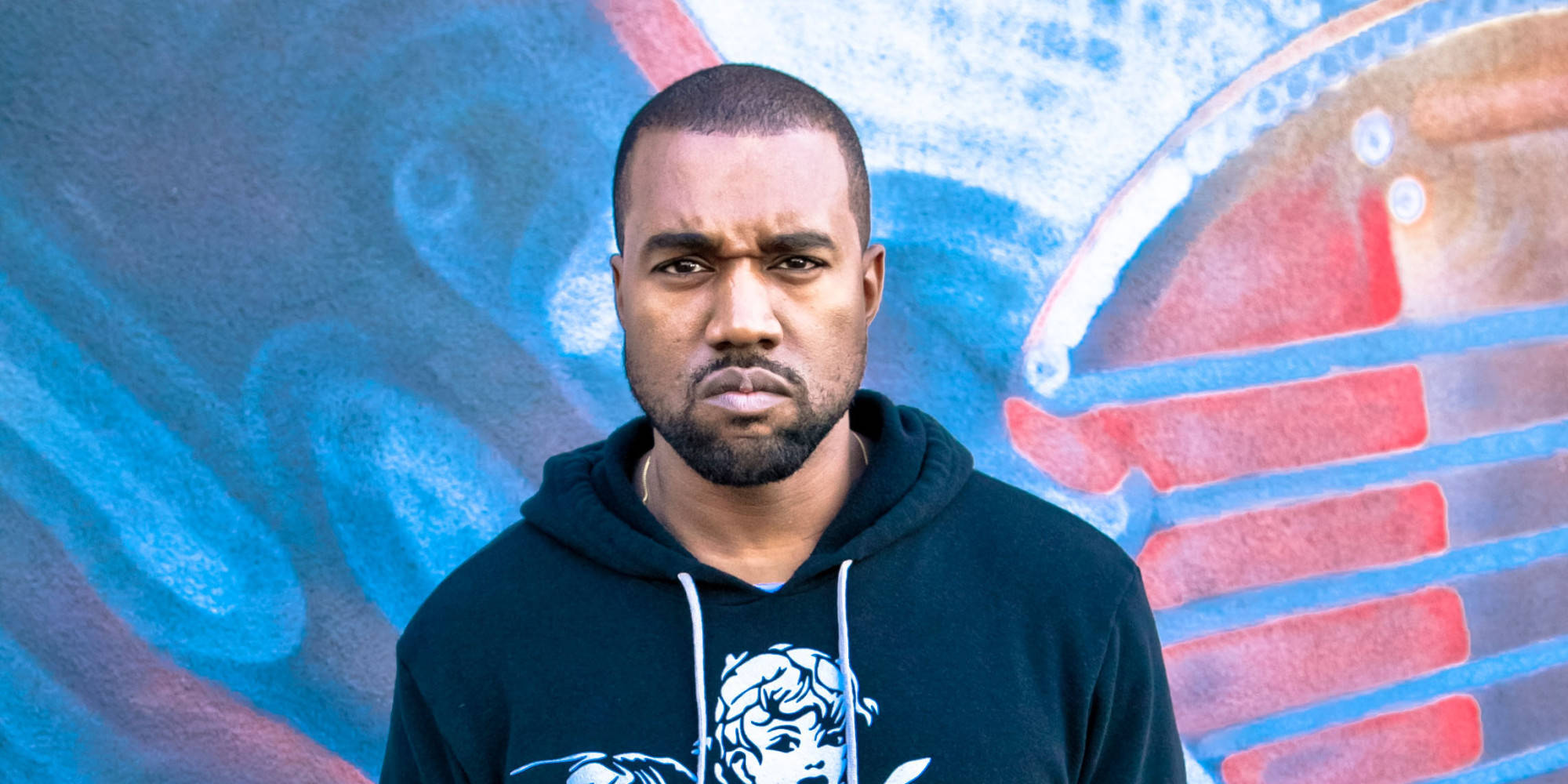 Kanye 桌布 壁紙 — 一名身穿黑色連帽衫的男子站在充滿活力的藍色和紅色塗鴉牆前，表情強烈。