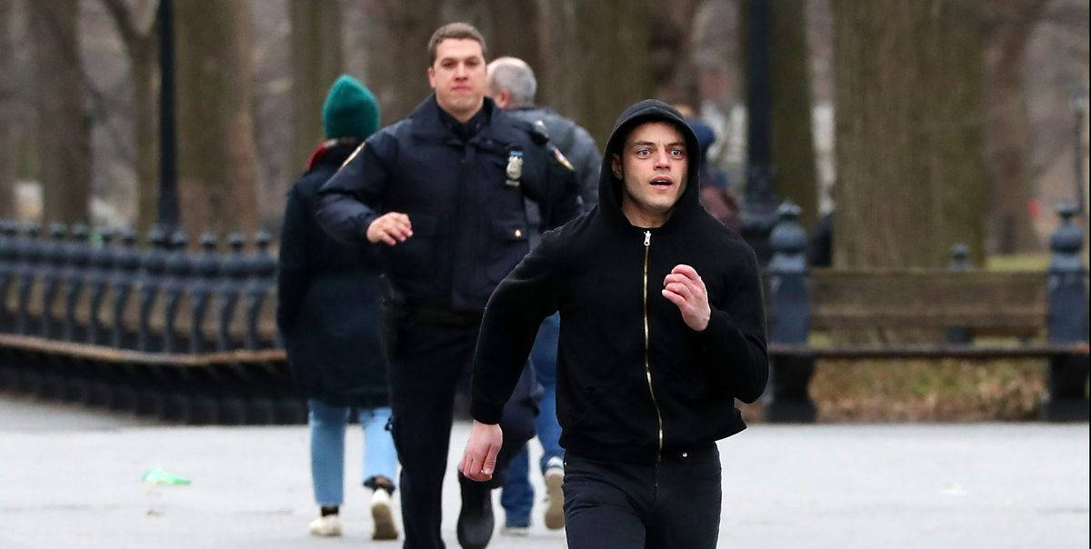 Rami Malek 桌布 壁紙 — 一名身穿黑色連帽衫的男子緊急跑過公園，一名警察和其他行人跟在後面。
