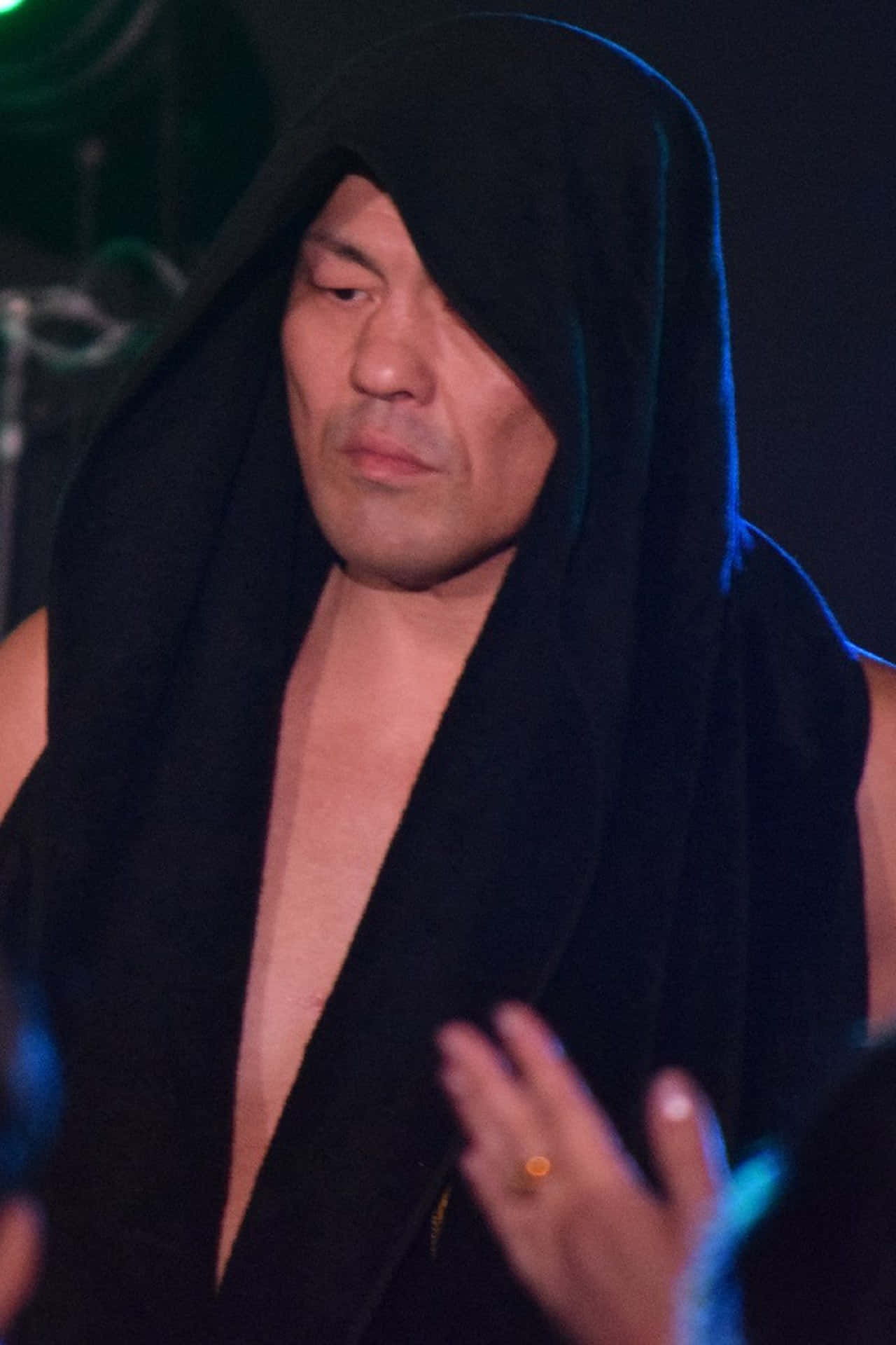 Aew 桌布 壁紙 — 一名身穿黑色連帽長袍、遮住一隻眼睛的男子，在昏暗的場地中被藍色舞台燈光照亮。