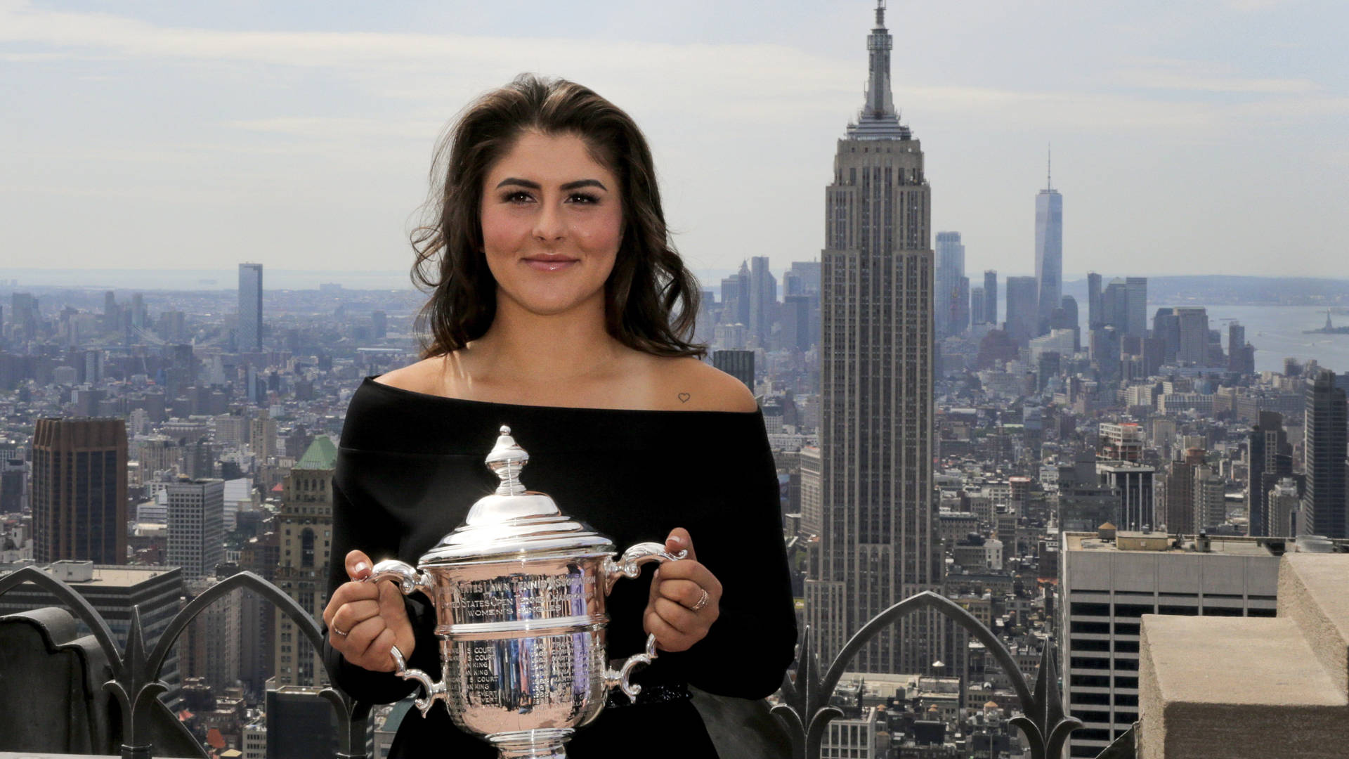 Bianca Andreescu 桌布 壁紙 — 一名身穿黑色連衣裙的女性，以紐約市標誌性的天際線為背景，手持閃亮的銀色獎盃。