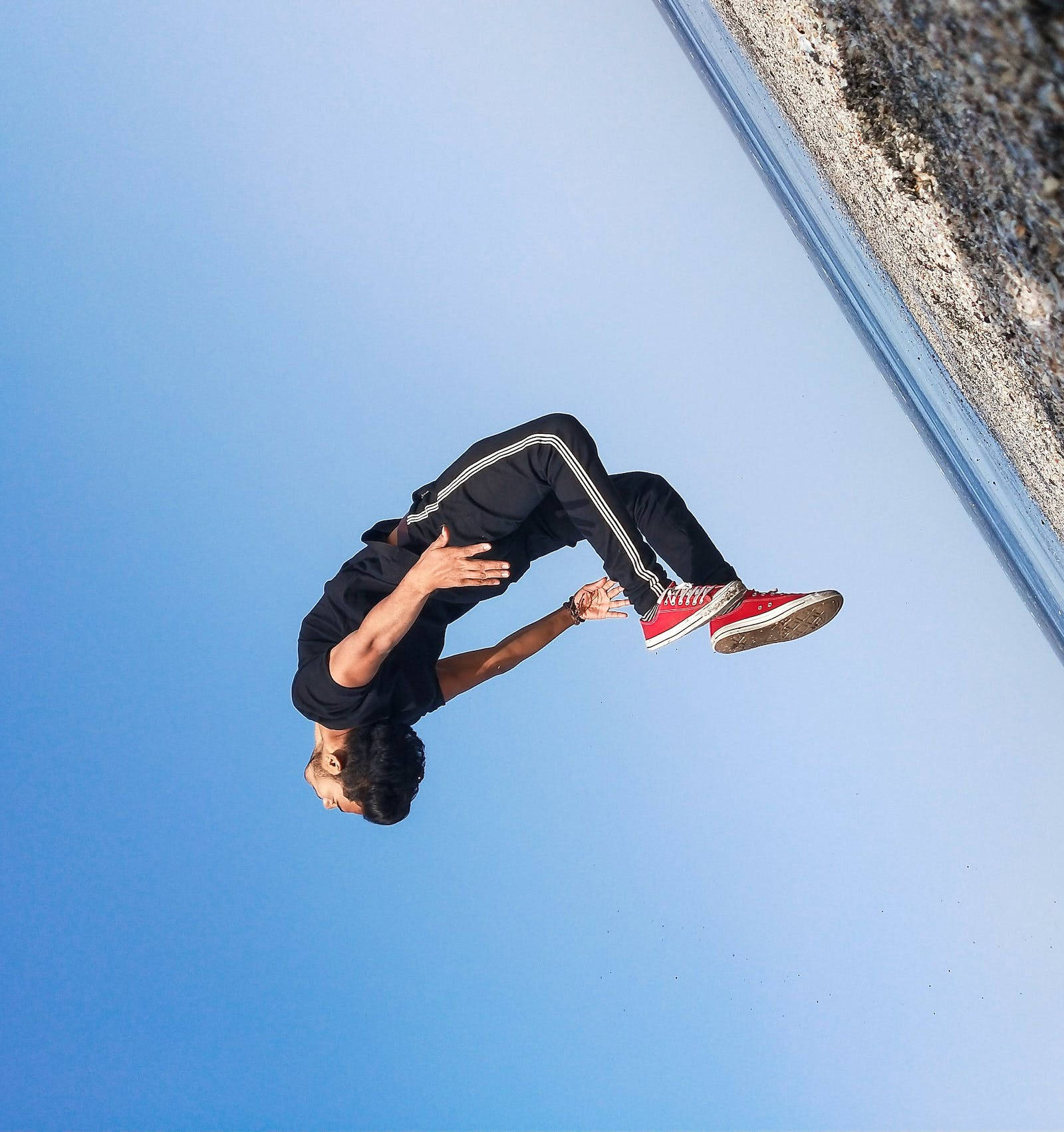 Jumping 桌布 壁紙 — 一名身穿黑色運動服和紅色運動鞋的年輕男子，在岩石海岸線附近清澈的藍天下做出動態的空中翻騰動作。