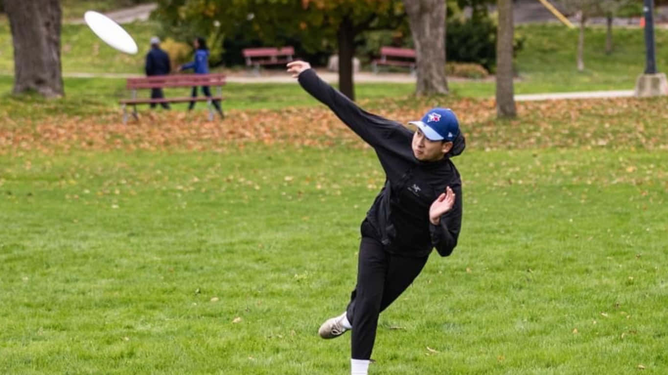 1366x768 Frisbee 桌布 壁紙 — 一名身穿黑色運動服的人將白色飛盤扔過一個散落著秋葉的草地公園。