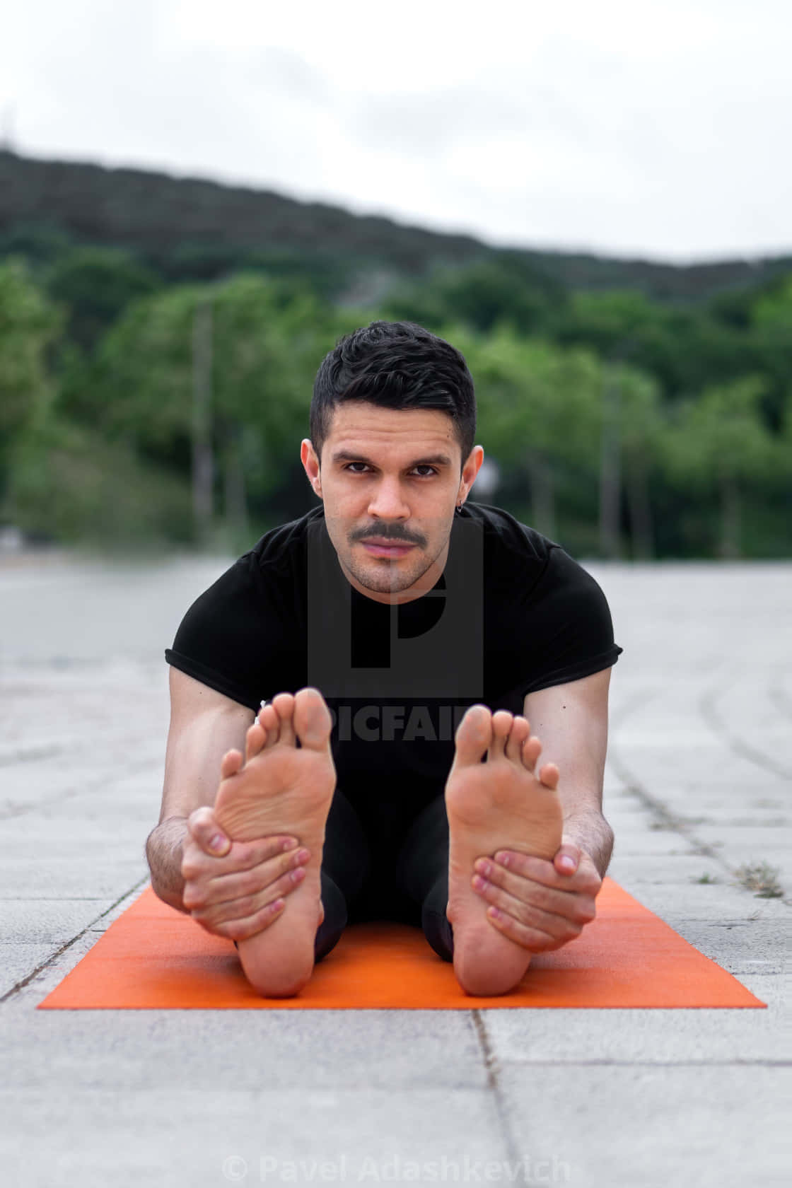 Yoga 桌布 壁紙 — 一名身穿黑色運動服的男士在戶外的橙色墊子上進行坐姿前屈伸展，背景有綠樹和山丘。