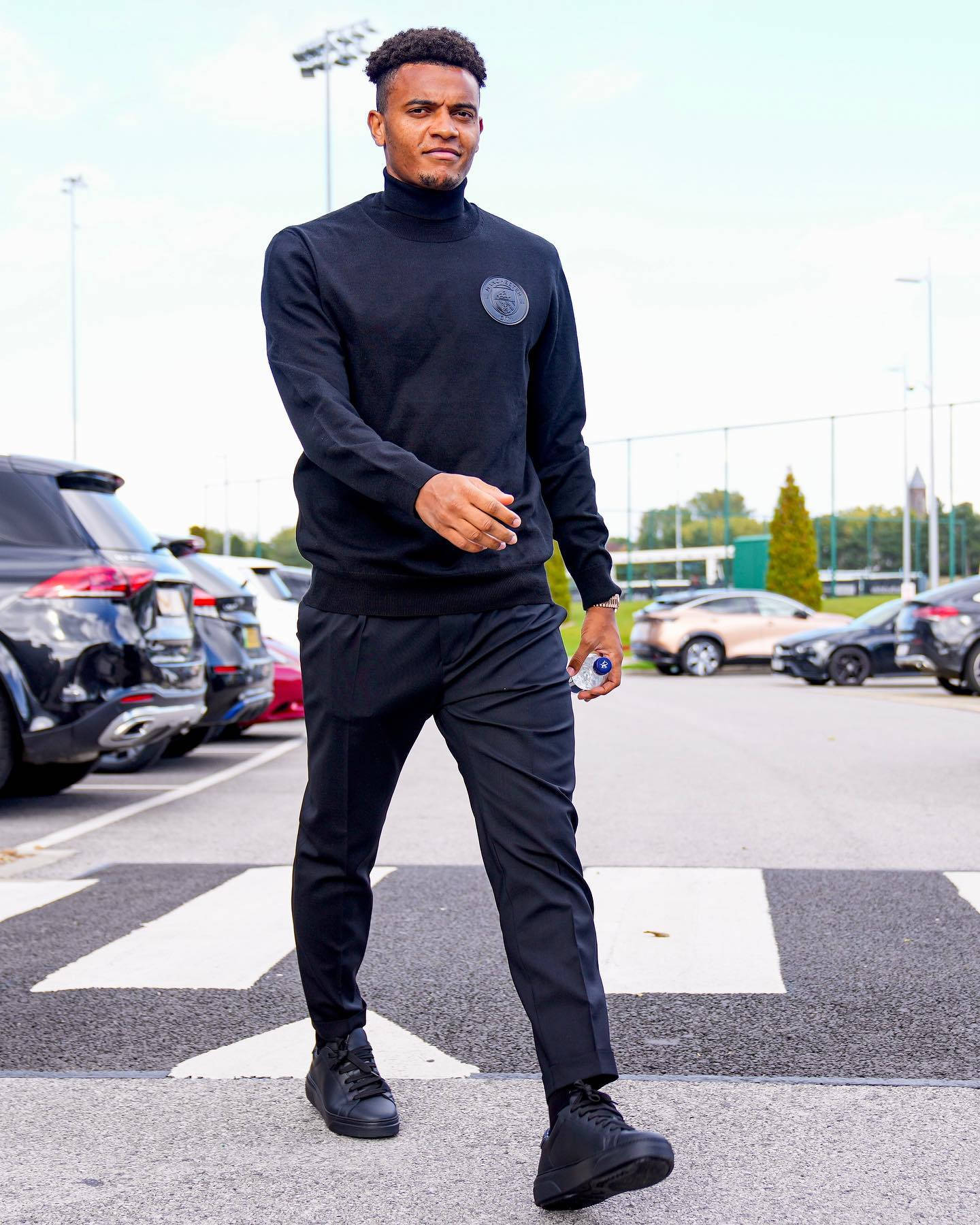 Celebrity 桌布 壁紙 — 一名身穿黑色運動服的男子在明亮的日光下自信地站在停車場的斑馬線上，手持水壺。