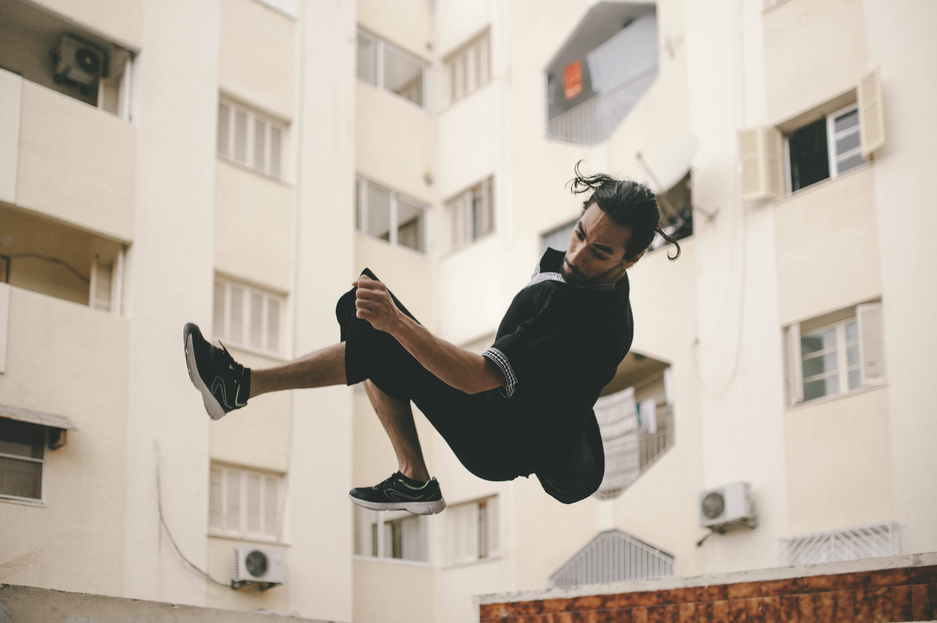 Parkour 桌布 壁紙 — 一名身穿黑色運動服的男子在米色公寓大樓外牆前騰空跳起，大樓有窗戶和空調機。