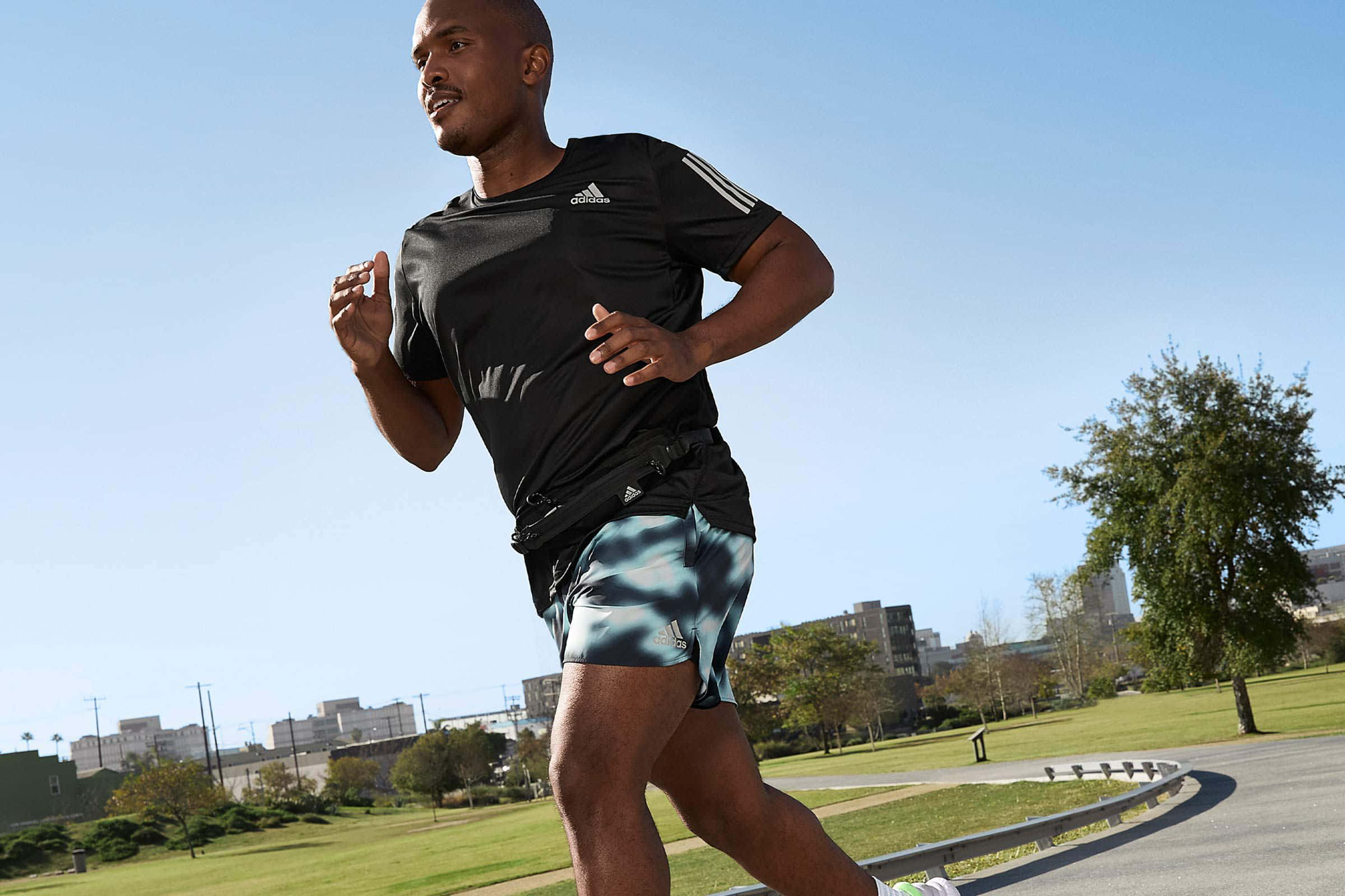 Running 桌布 壁紙 — 一名身穿黑色運動服的男子在陽光明媚的城市公園中，沿著鋪砌的小徑奔跑，周圍有綠草和遠處的建築物，天空清澈湛藍。