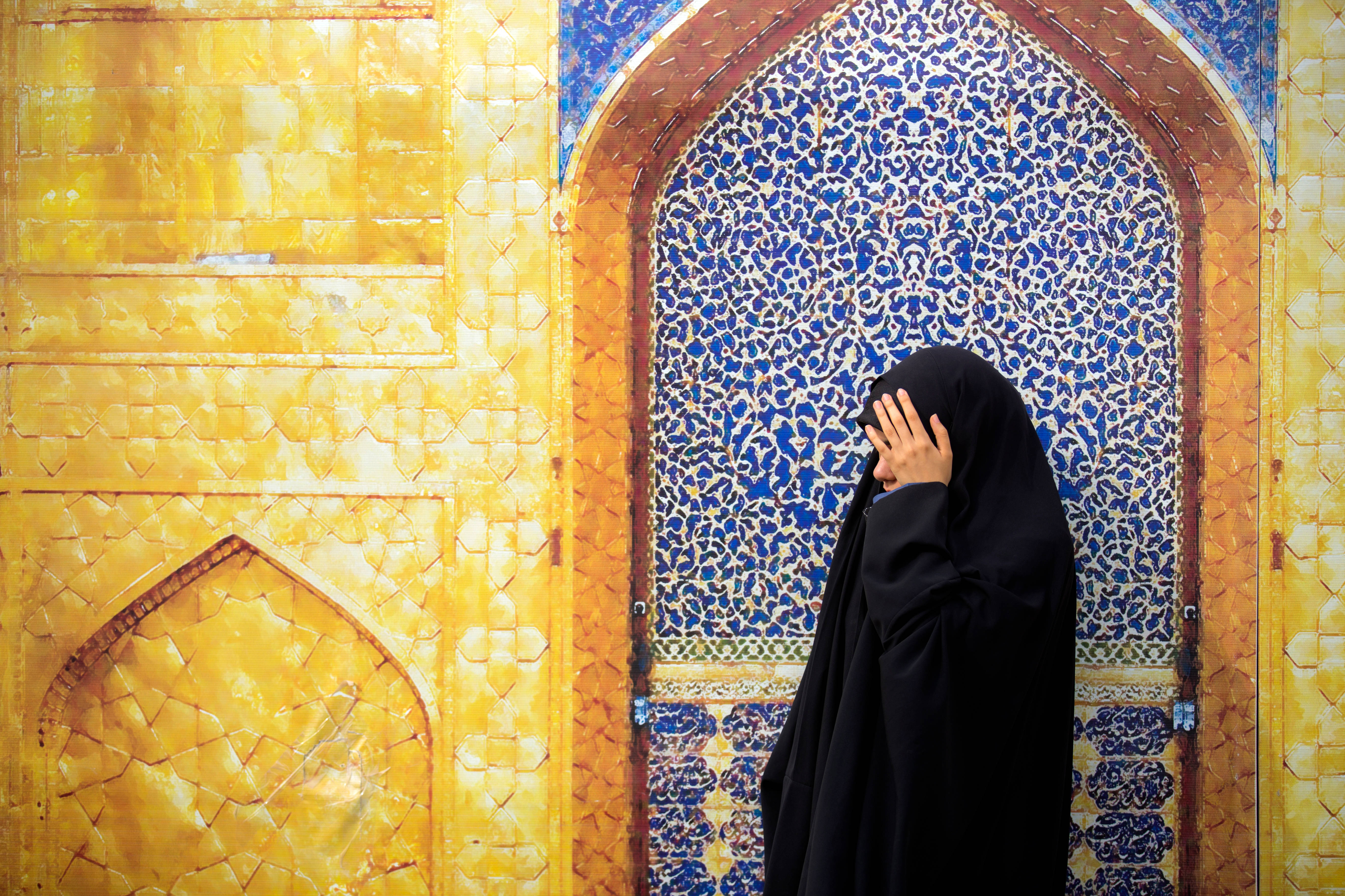 Best Islamic 桌布 壁紙 — 一名身穿黑色長袍的女子站在華麗的藍金色伊斯蘭瓷磚拱門前，手撫臉頰，靜默沉思。