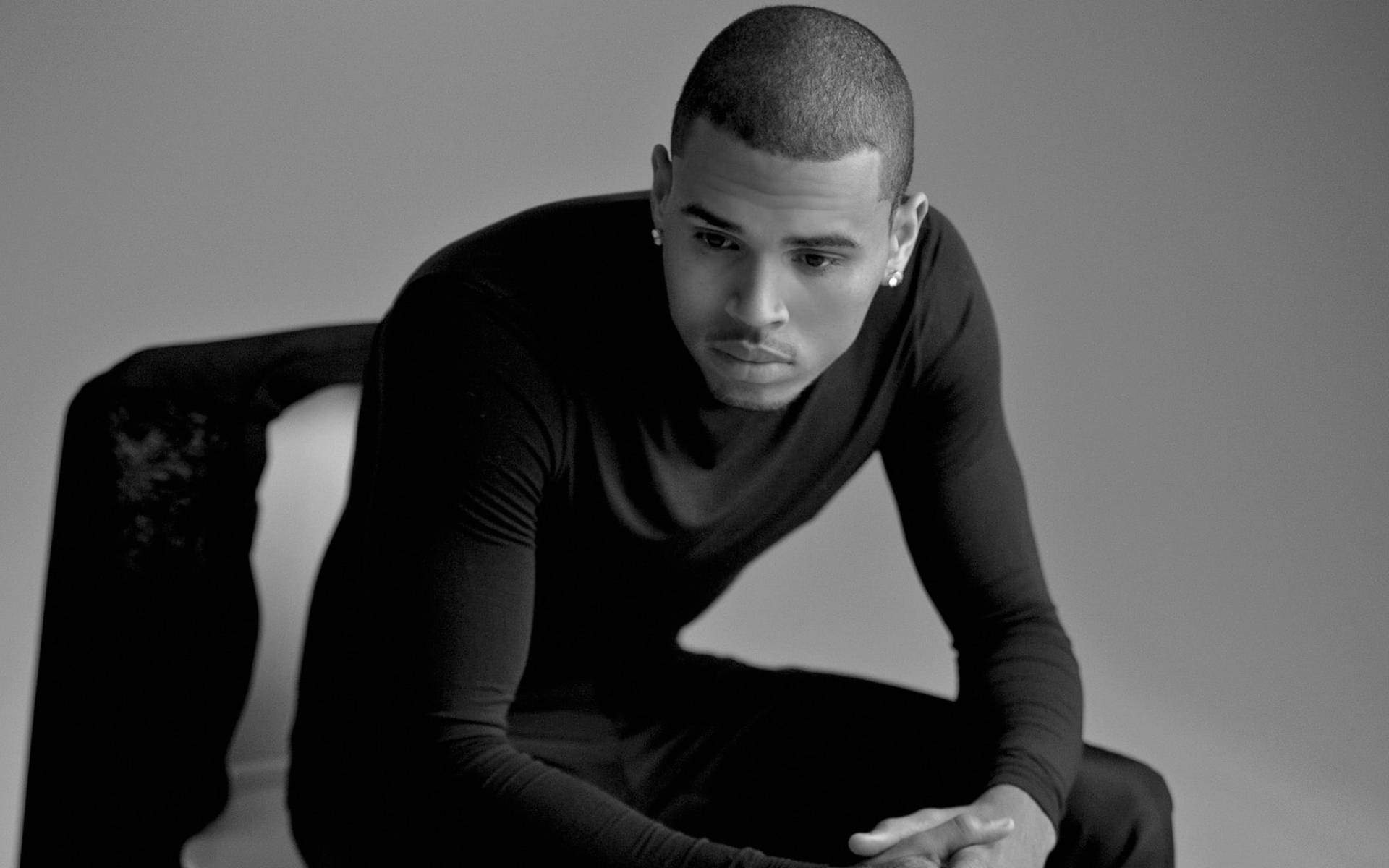 Chris Brown 桌布 壁紙 — 一名身穿黑色長袖襯衫的男子坐在椅子上，表情嚴肅，身處簡約風格的攝影棚環境中。