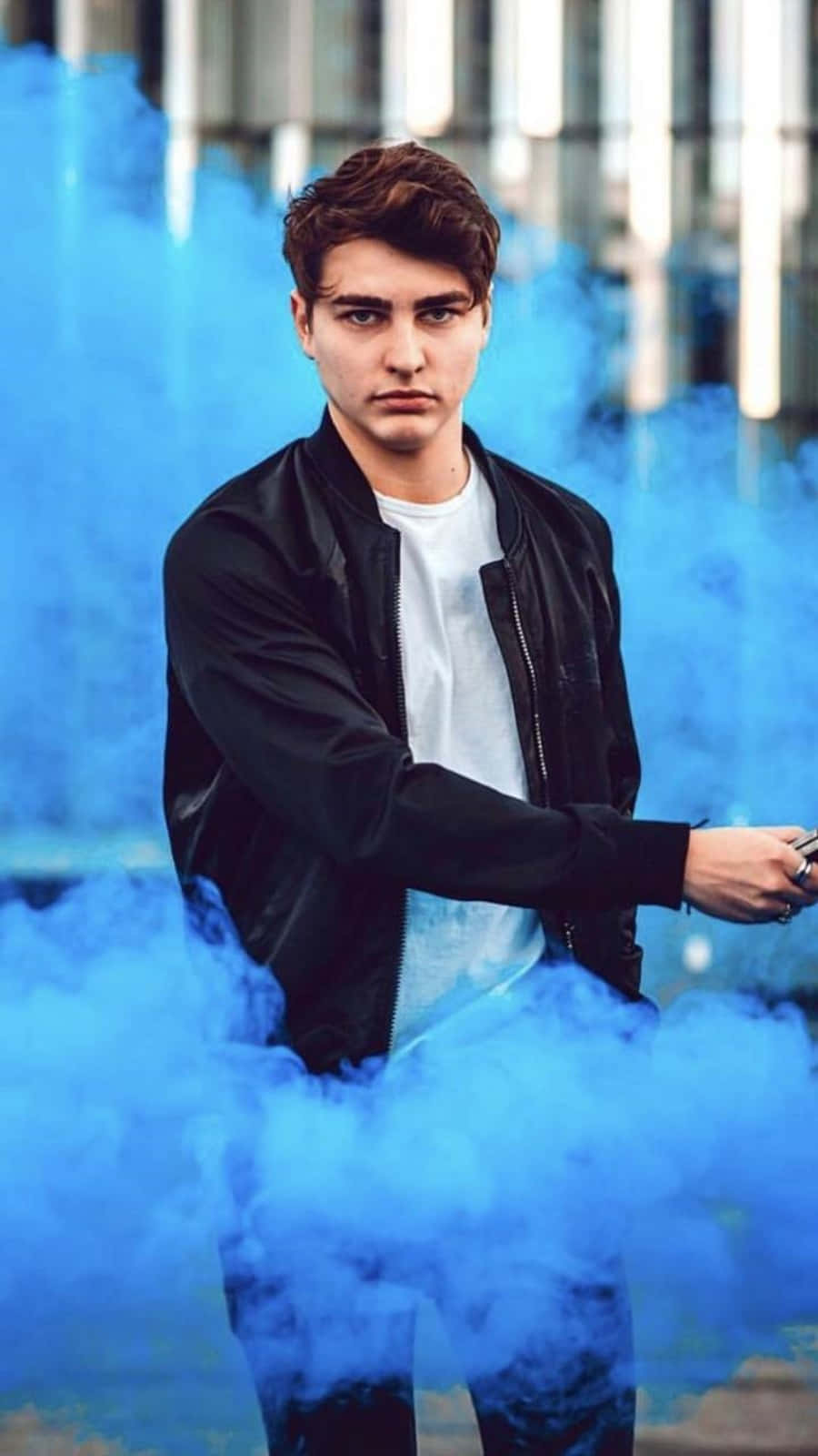 Colby Brock 桌布 壁紙 — 一名身穿黑色飛行員夾克的年輕男子站在旋轉的藍色煙霧中，背景是模糊的現代建築。