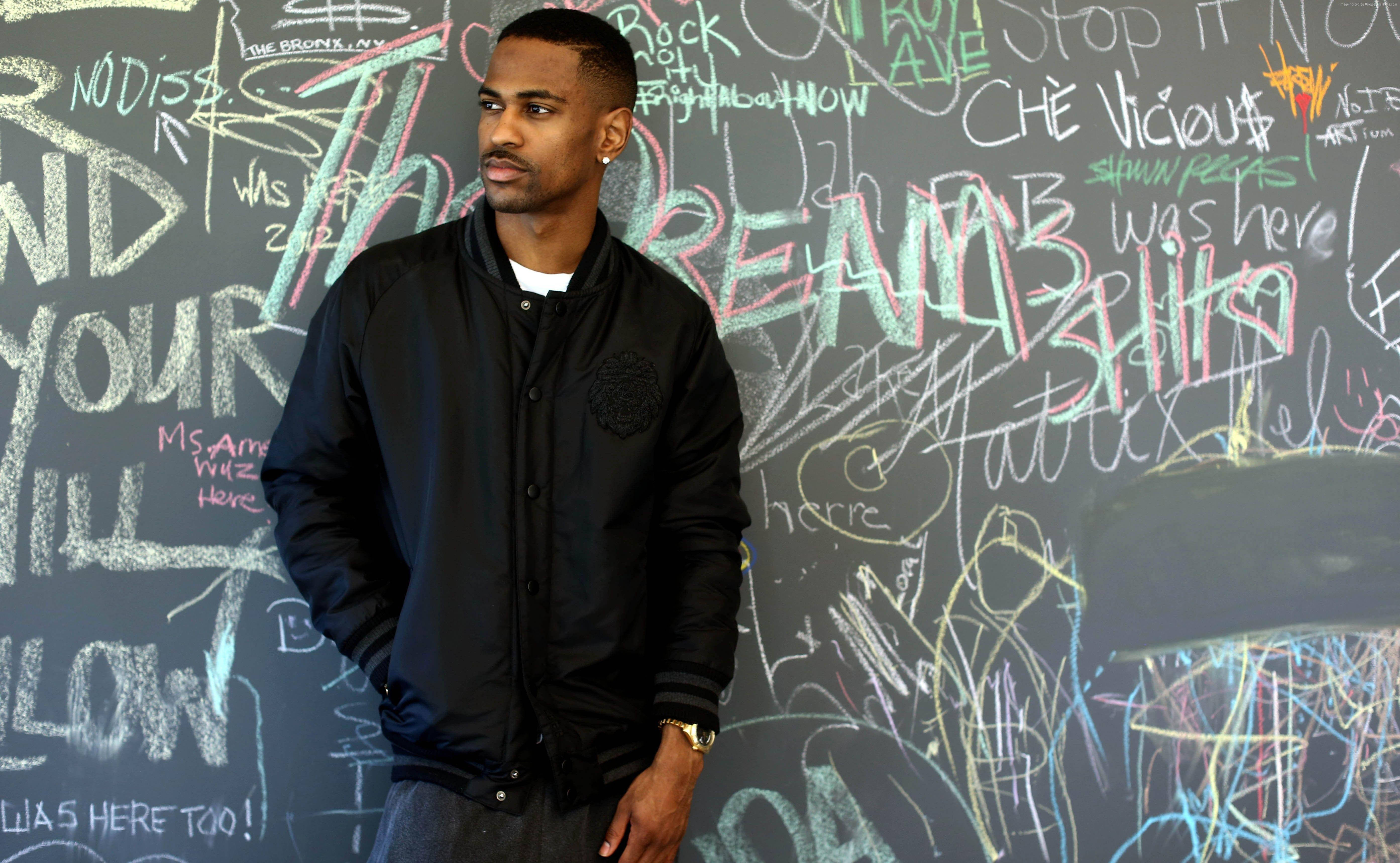 Big Sean 桌布 壁紙 — 一名身穿黑色飛行員夾克的男子自信地站在一面充滿色彩繽紛塗鴉標籤和訊息的牆前。