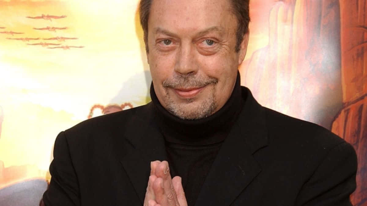Tim Curry 桌布 壁紙 — 一名身穿黑色高領毛衣和西裝外套的男子，在色彩繽紛的動畫背景前雙手合十，溫暖地微笑。