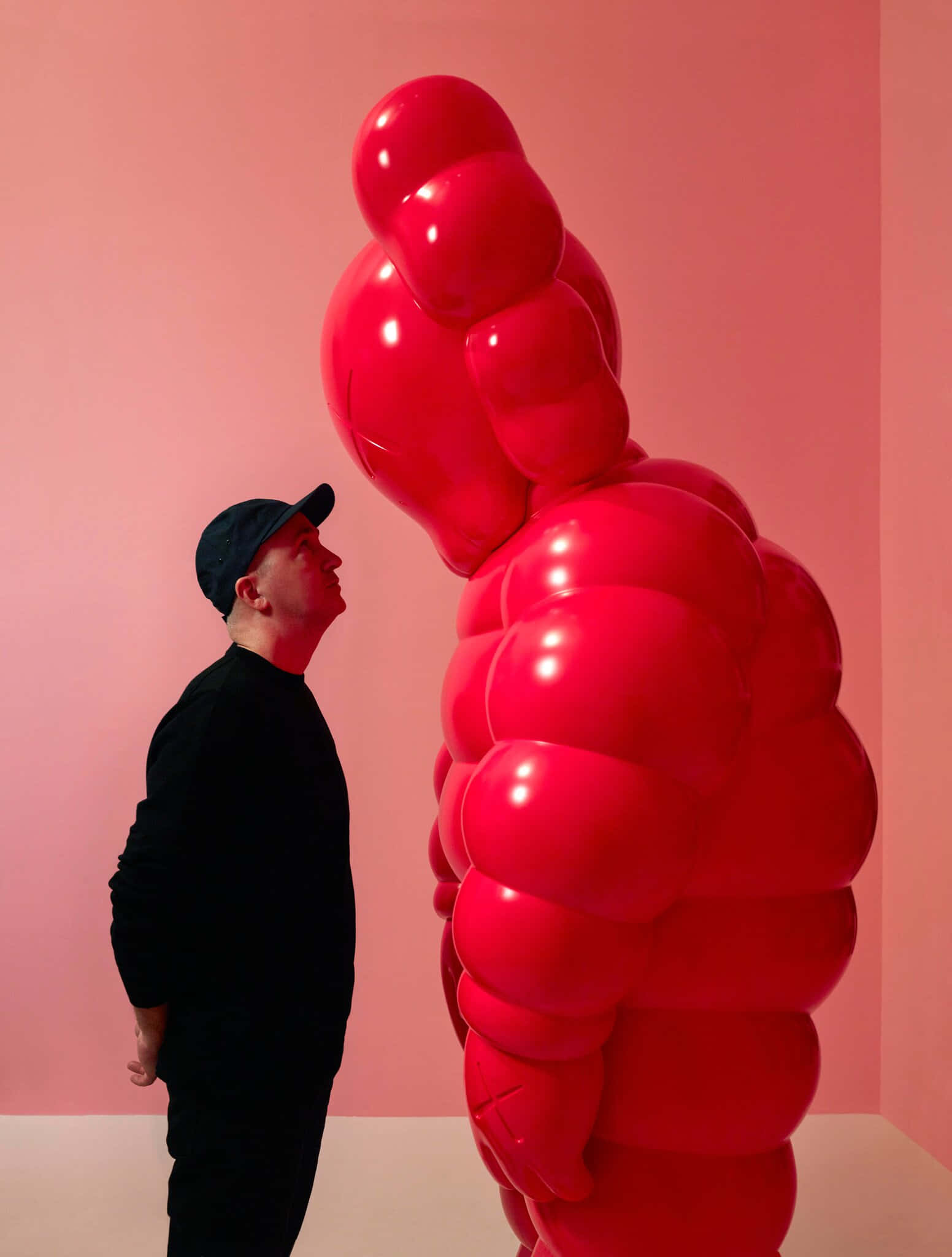 KAWS 桌布 壁紙 — 一名身穿黑衣的男子敬畏地站在一座高聳的亮紅色氣球雕塑旁，背景是柔和的粉紅色牆壁。