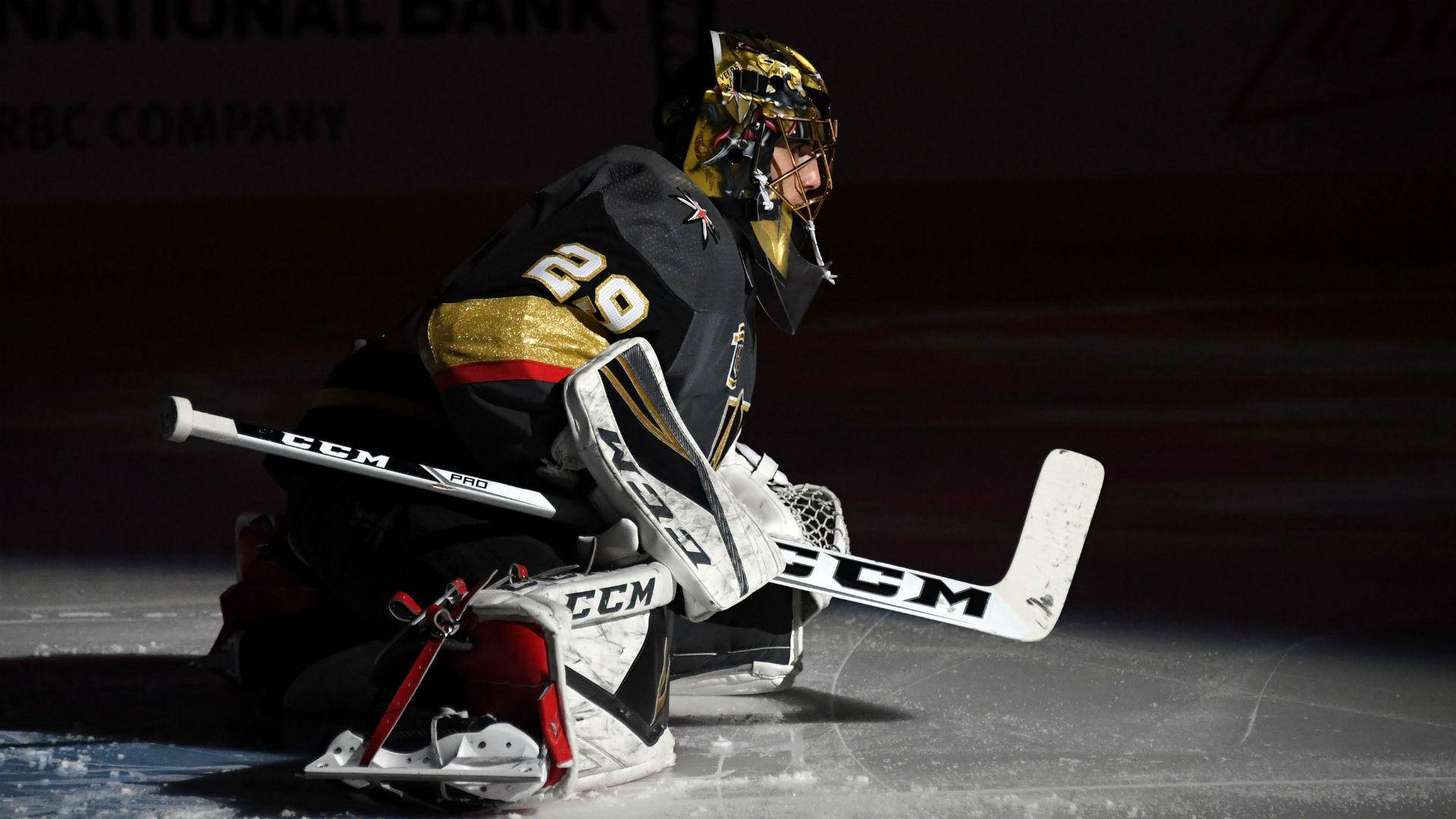 hockey 桌布 壁紙 — 一名身穿黑金色裝備的冰球守門員在場館燈光下低蹲在冰上，手持球桿。