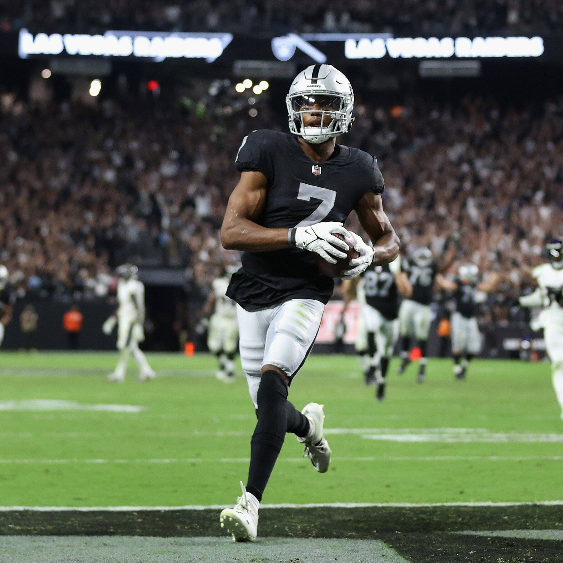 zay jones 桌布 壁紙 — 一名身穿黑銀色球衣的拉斯維加斯突襲者美式足球員，在明亮的燈光下，帶著球衝過擠滿觀眾的體育場。