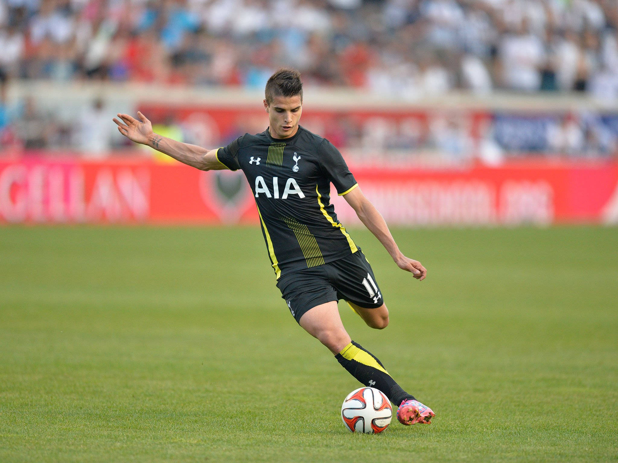 erik lamela 桌布 壁紙 — 一名身穿黑黃色球衣的足球運動員在充滿活力的綠色球場上運球，球為白色和紅色，背景有模糊的觀眾。