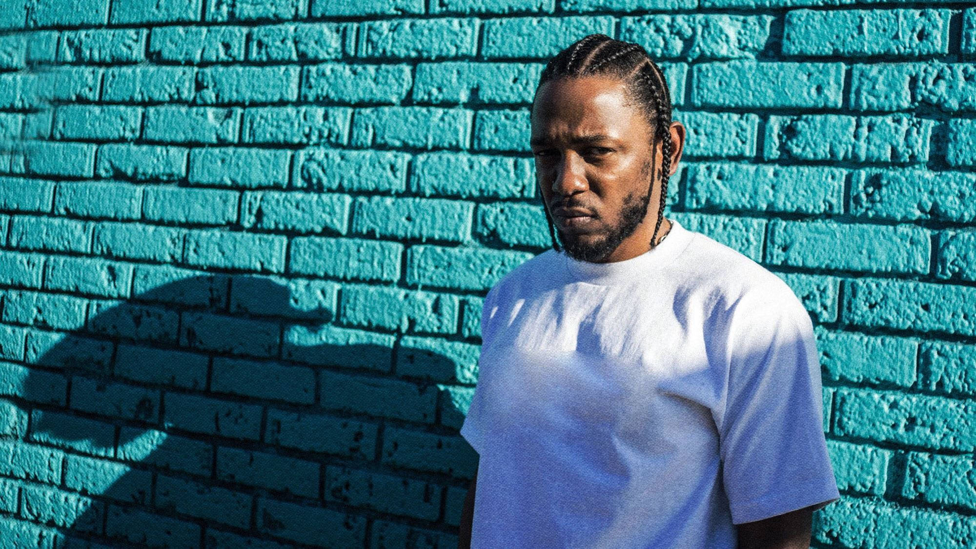 Kendrick 桌布 壁紙 — 一名辮子男子站在鮮豔的青綠色磚牆前，在明亮的陽光下投射出清晰的陰影。