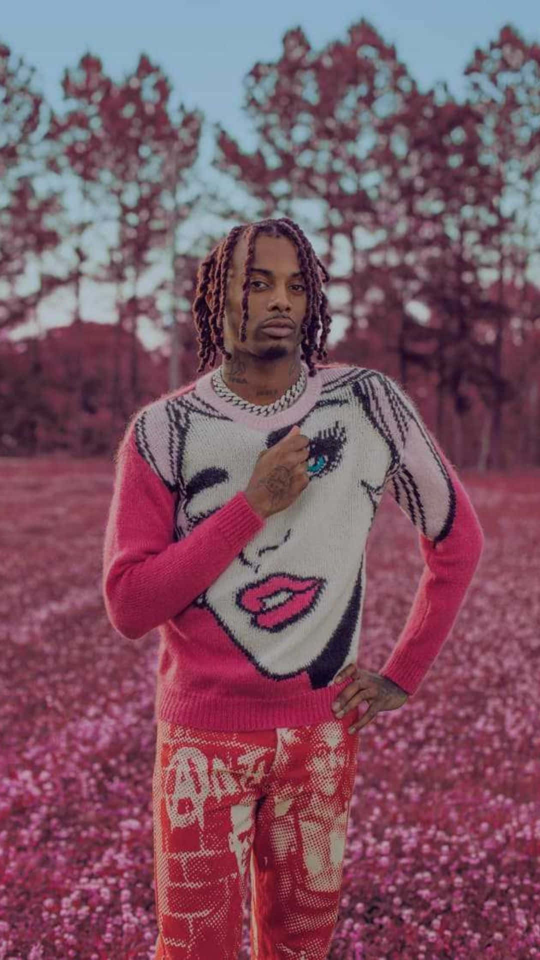 Playboi Carti 桌布 壁紙 — 一名辮子男子自信地站在粉紅色花田中，身穿帶有風格化臉部圖案的粉紅色毛衣和配套的圖案長褲，背景是暮色天空。