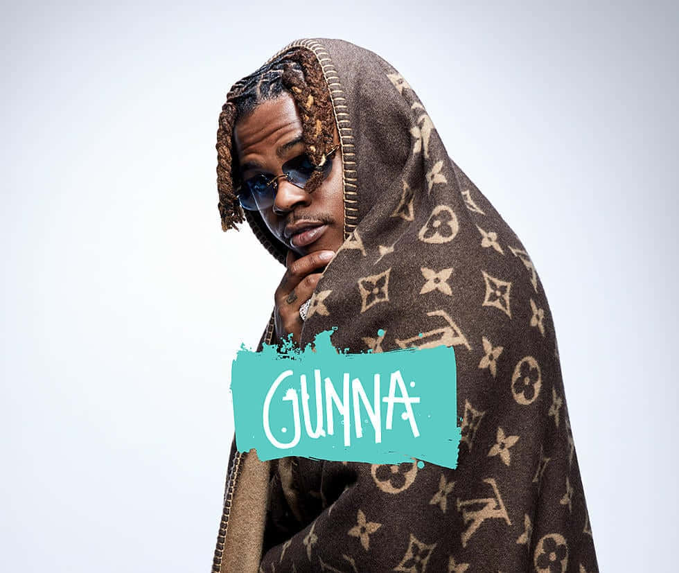 Gunna 桌布 壁紙 — 一名辮子頭髮、戴太陽眼鏡的男子，披著棕色 Louis Vuitton 字母圖案毛毯，在純白背景前沉思地擺姿勢。