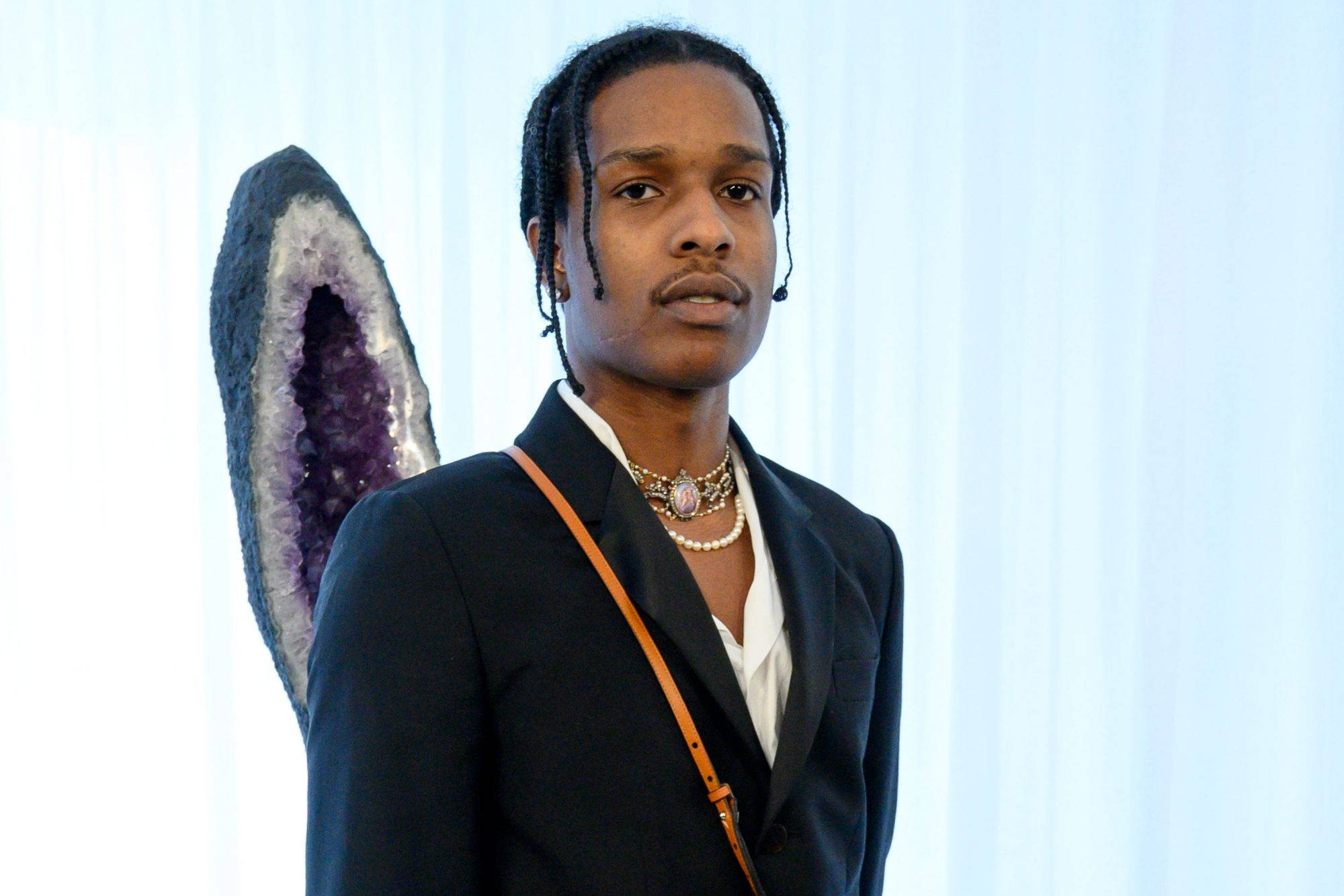 rocky 桌布 壁紙 — 一名辮子頭髮、戴著多層項鍊的男子站在一個大型紫水晶晶洞旁邊，背景是淺藍色窗簾。