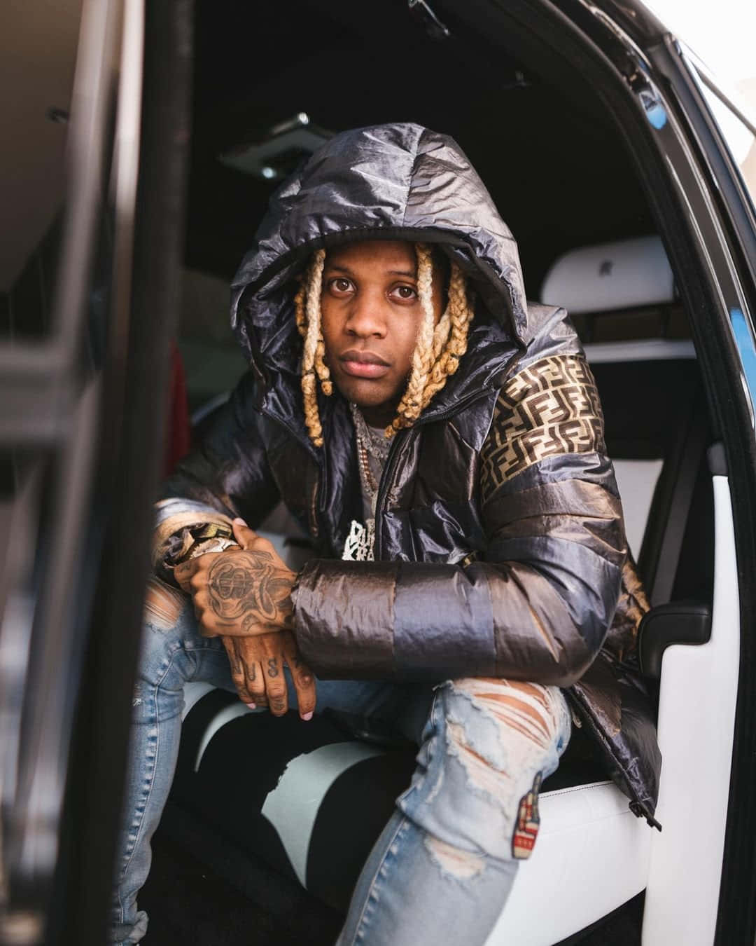 Lil Durk 桌布 壁紙 — 一名辮髮且有刺青的男子坐在車內，身穿帶有金色標誌袖子的閃亮黑色羽絨外套。
