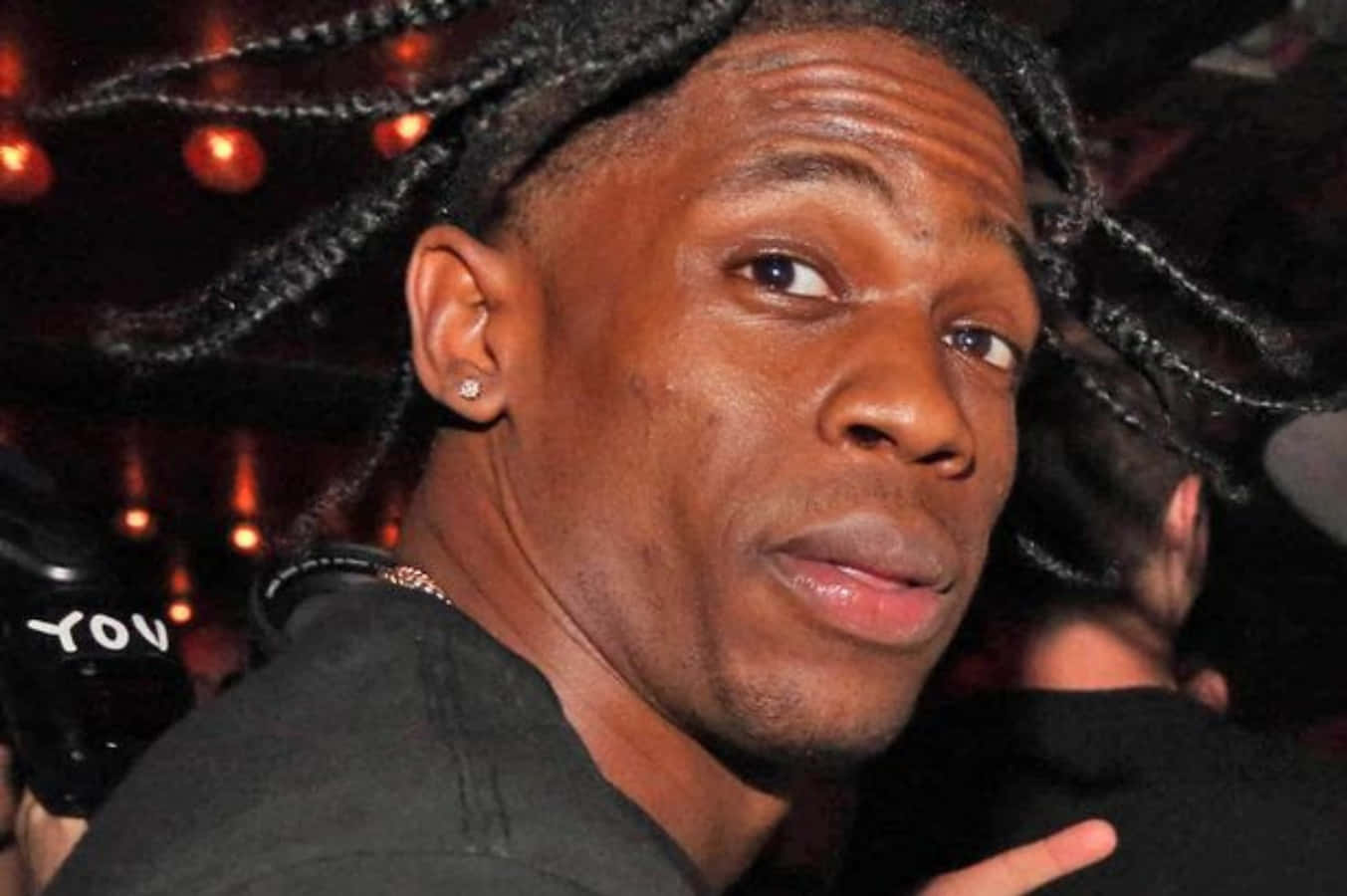 Travis Scott Funny 桌布 壁紙 — 一名辮髮男子戴著鑽石耳環，在燈光昏暗、溫暖光線的俱樂部中直視鏡頭的近距離肖像。