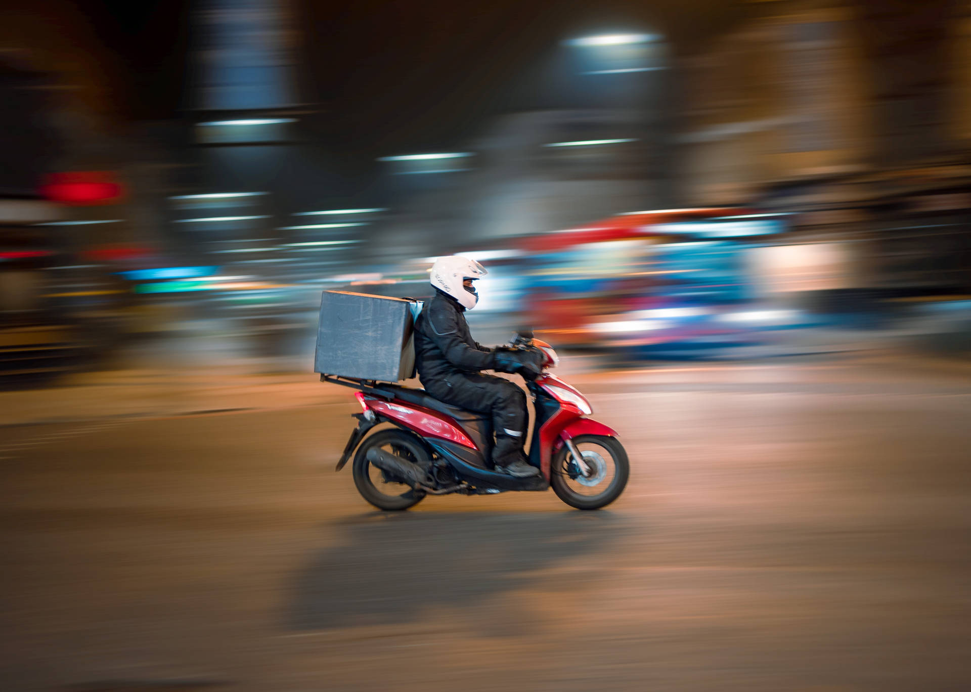  壁紙 — 一名送貨員騎著紅色機車，在模糊的城市夜晚街道上疾馳，攜帶一個大黑箱。